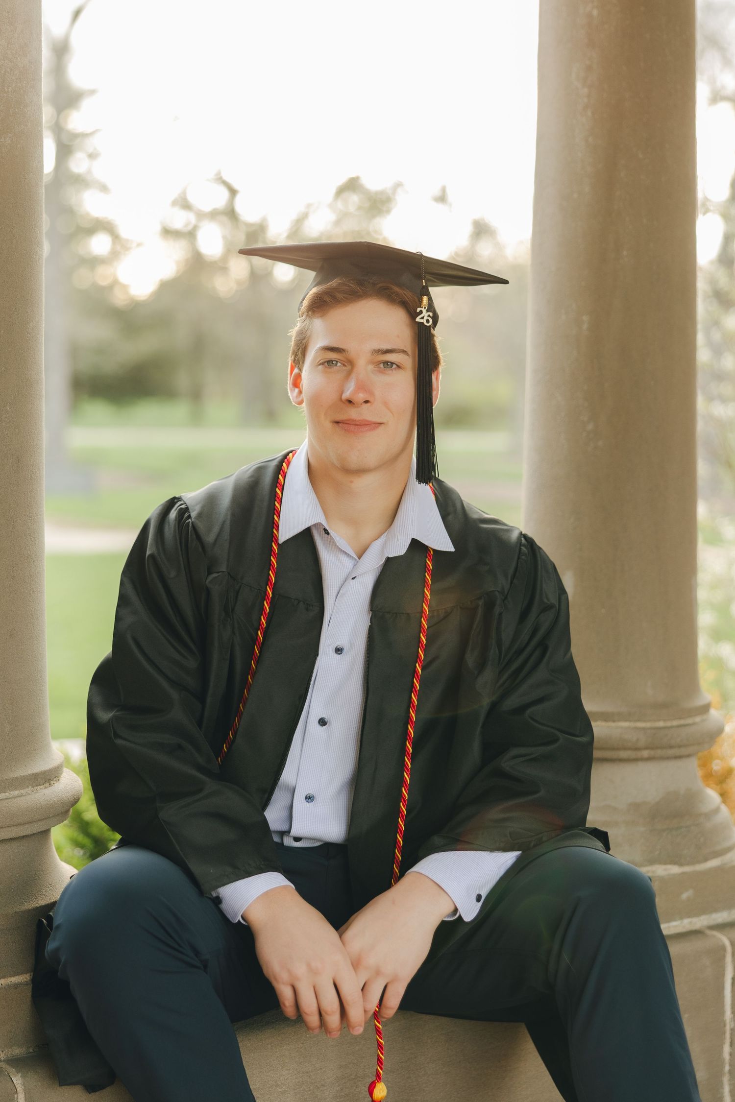 Young man in graduation cap and gown with honor cords sits between stone columns outdoors.