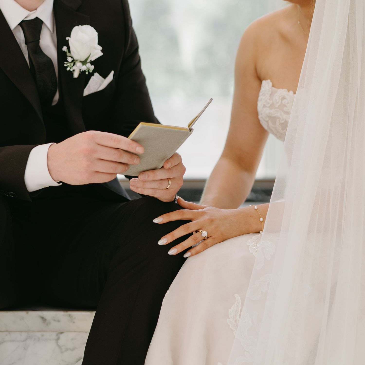 Groom reads wedding vows from a notebook while bride in white lace dress stands beside him during outdoor ceremony.