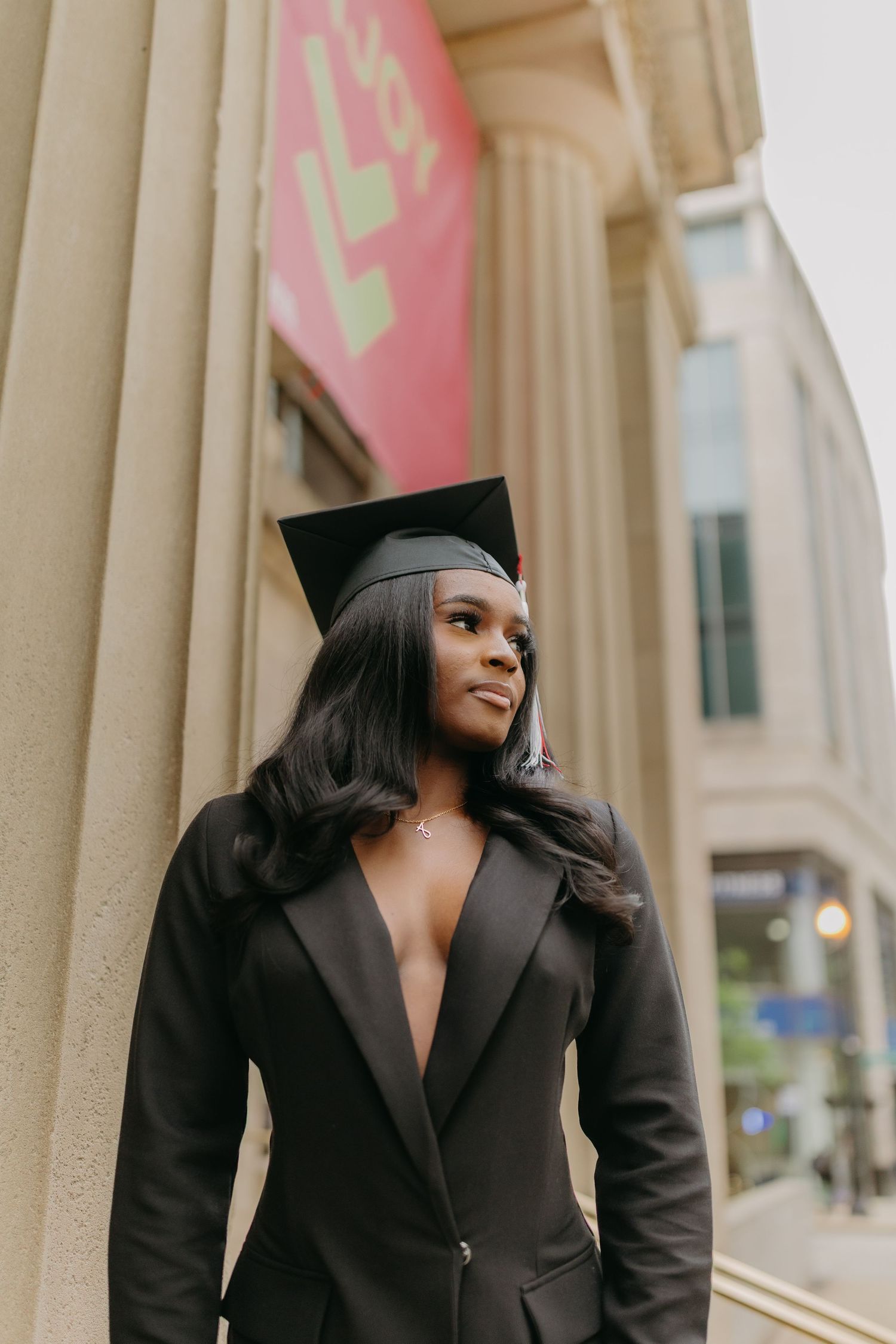 Graduate in black cap and gown poses confidently against architectural columns during graduation photoshoot.