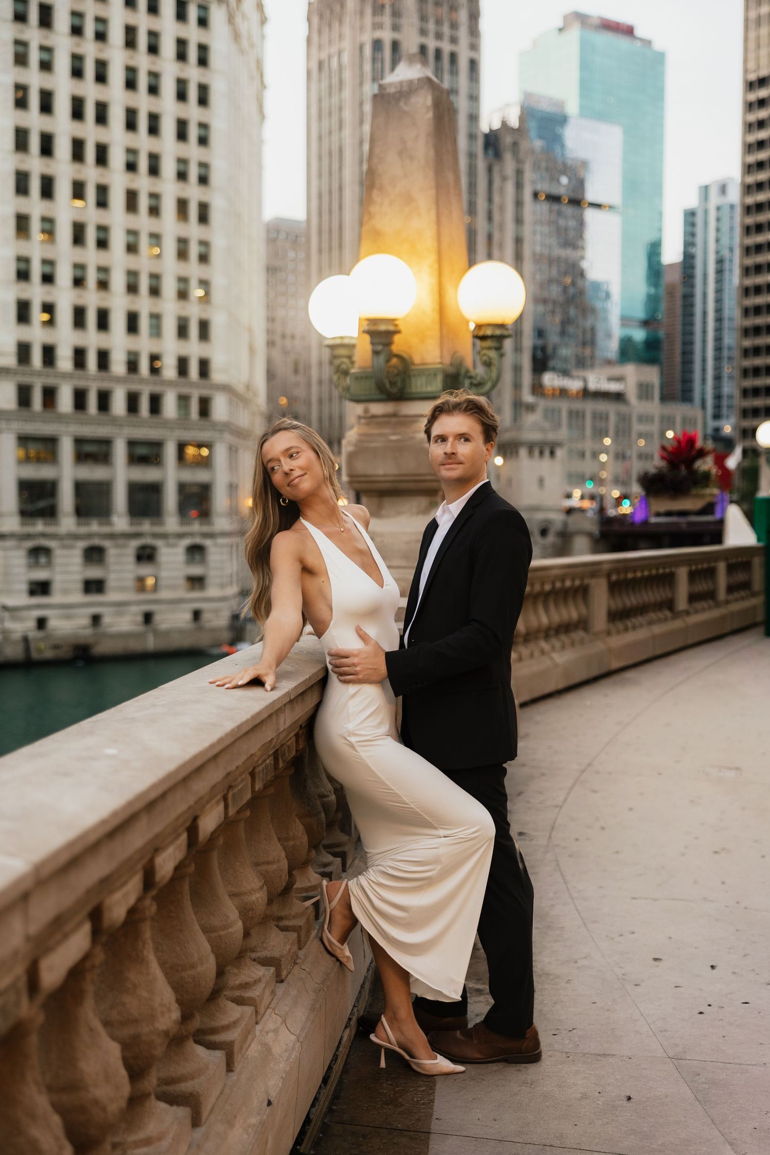 Elegant couple posing on Chicago Riverwalk bridge at dusk, woman in white gown, man in black suit, city skyline backdrop.