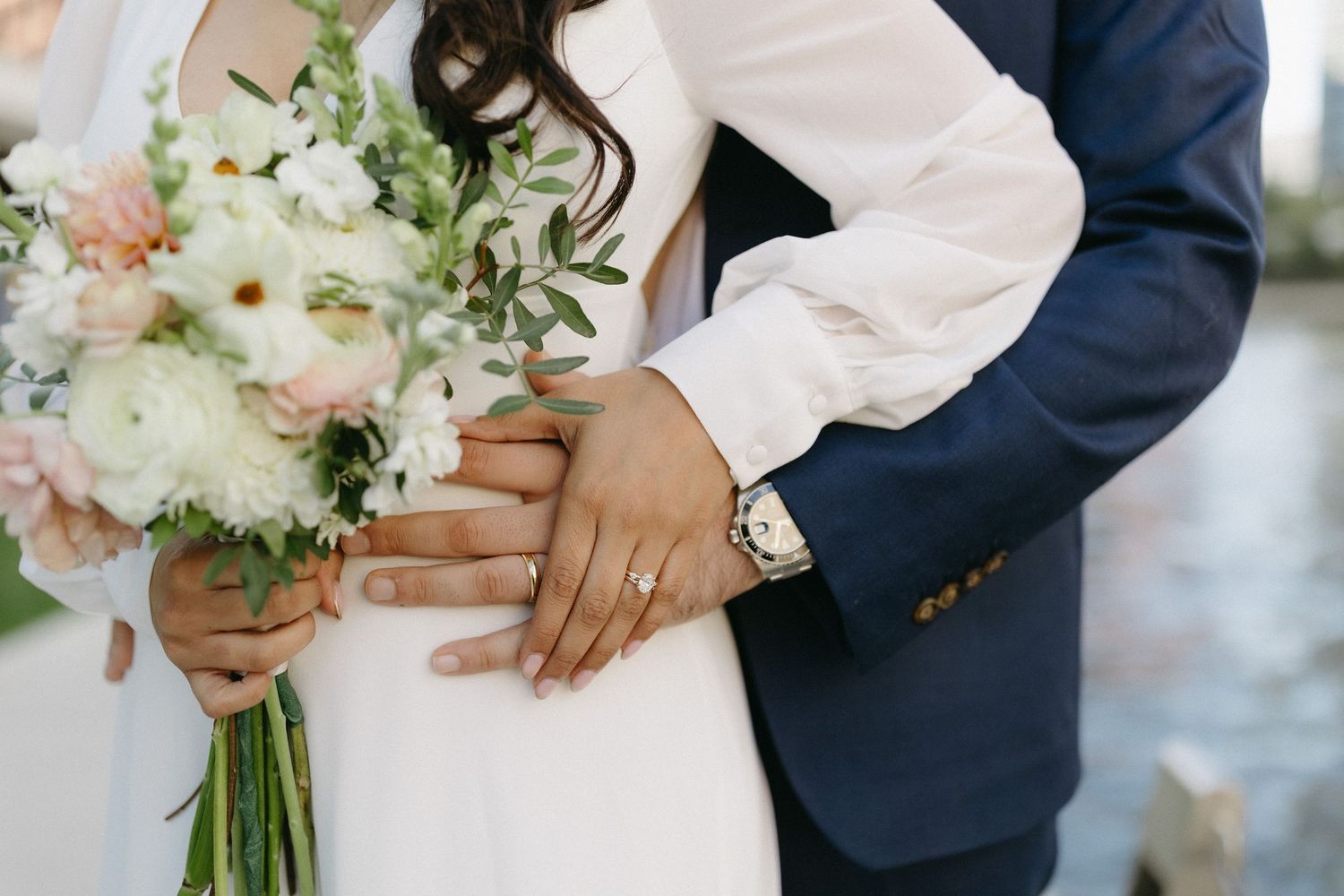 A wedding couple holds hands while showcasing a beautiful bouquet of white and blush flowers.