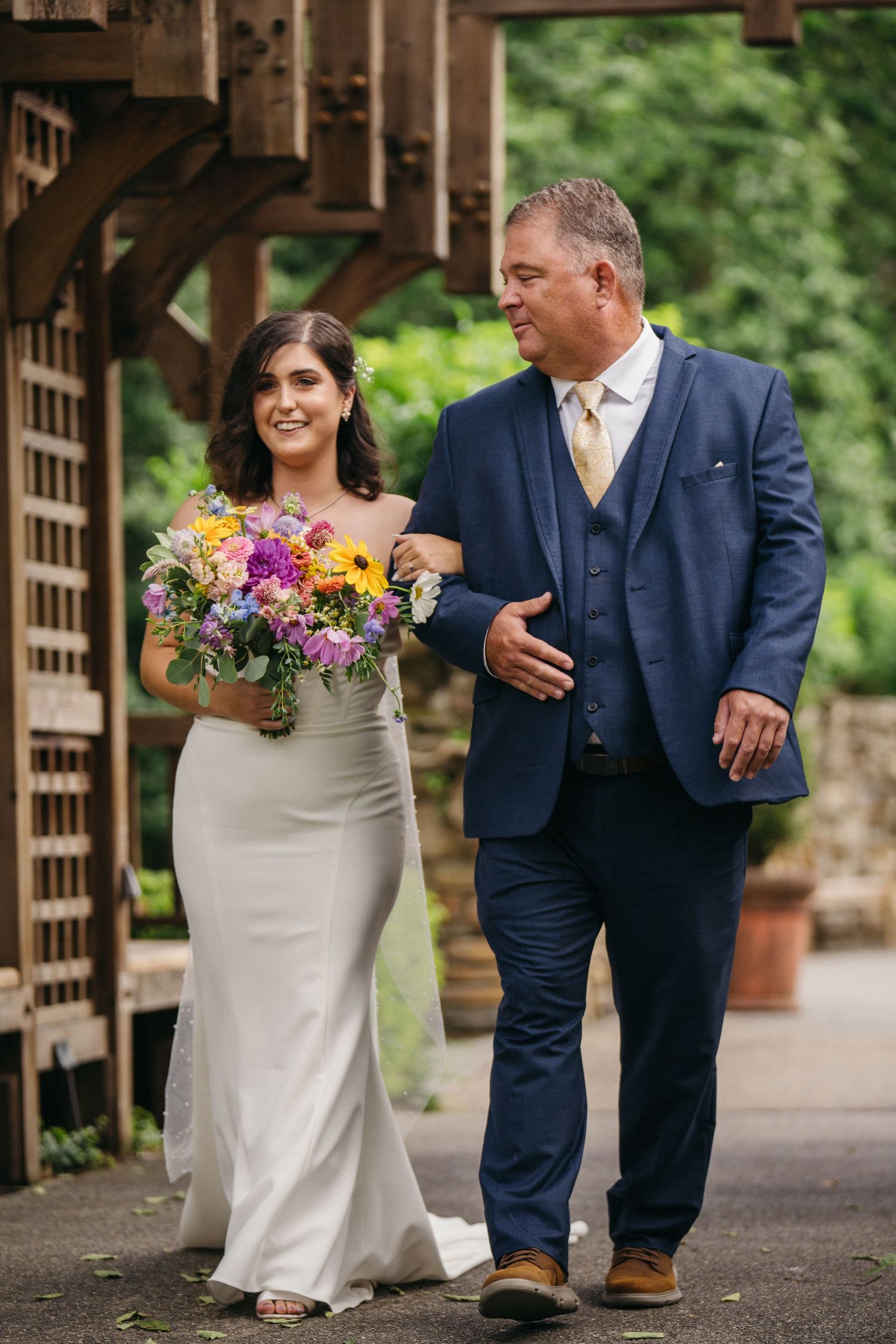 Bride in white gown holding colorful bouquet walks with father in blue suit at wedding.