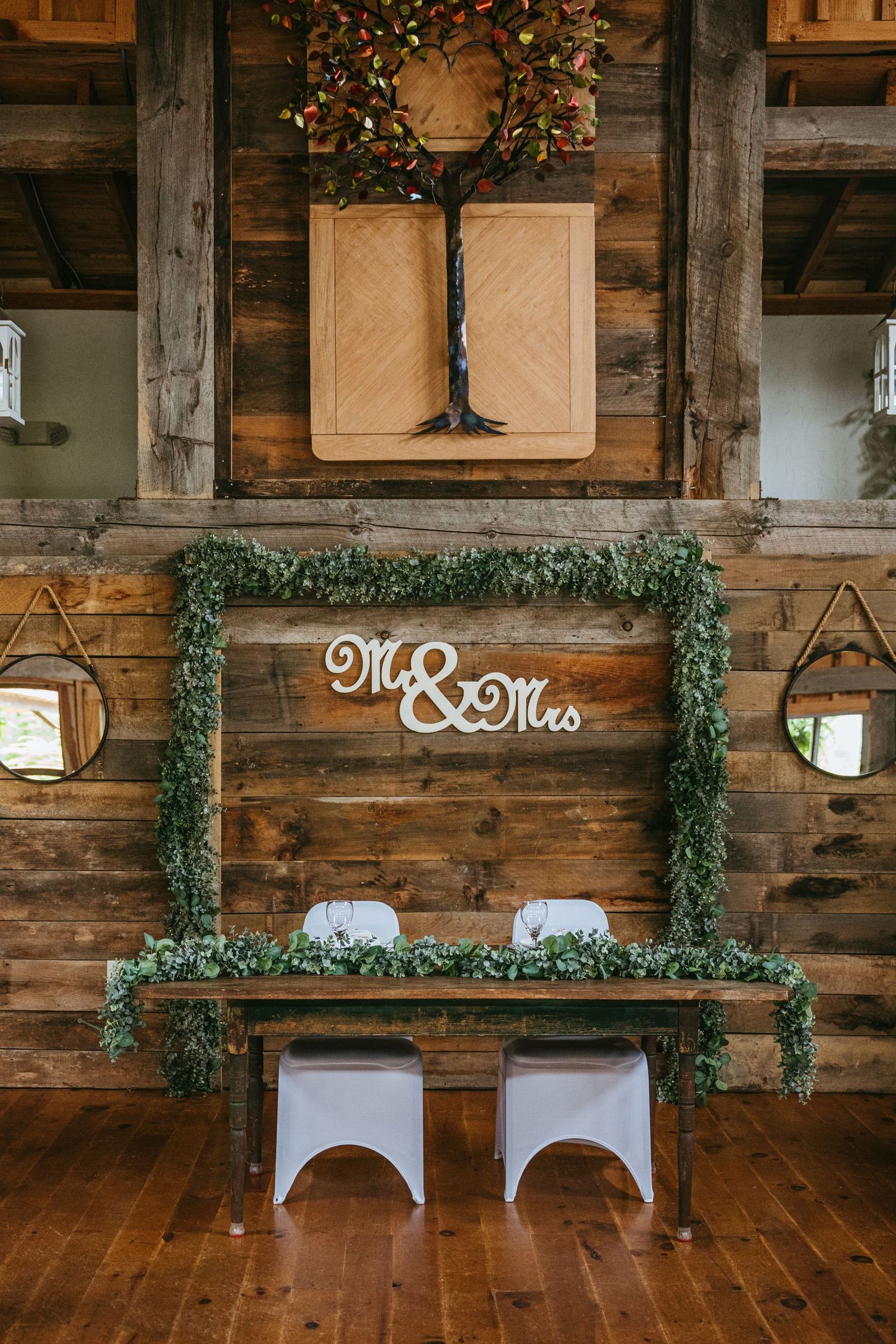 Rustic Mr. & Mrs. sweetheart table decorated with lush greenery garland in barn venue.