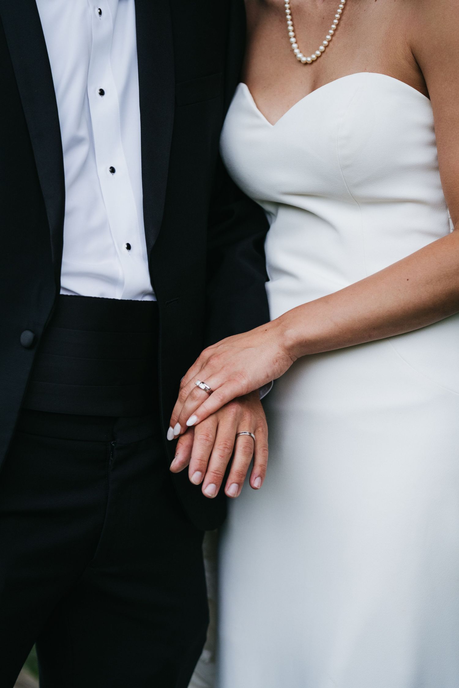 Bride and groom holding hands showing wedding rings, bride in strapless white gown with pearl necklace.