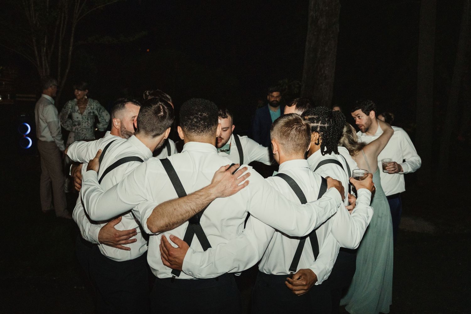 Groomsmen in white shirts and suspenders huddle together on the dance floor at a nighttime wedding reception.