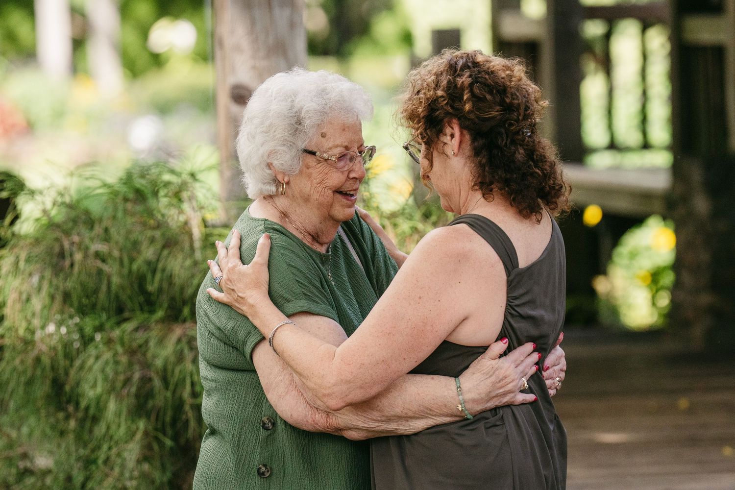 Two women sharing a warm embrace outdoors at a wedding celebration surrounded by greenery.