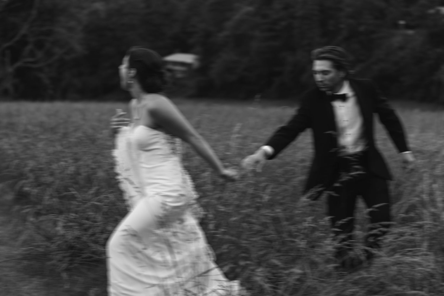 Black and white photo of bride and groom running hand in hand through a grassy field.