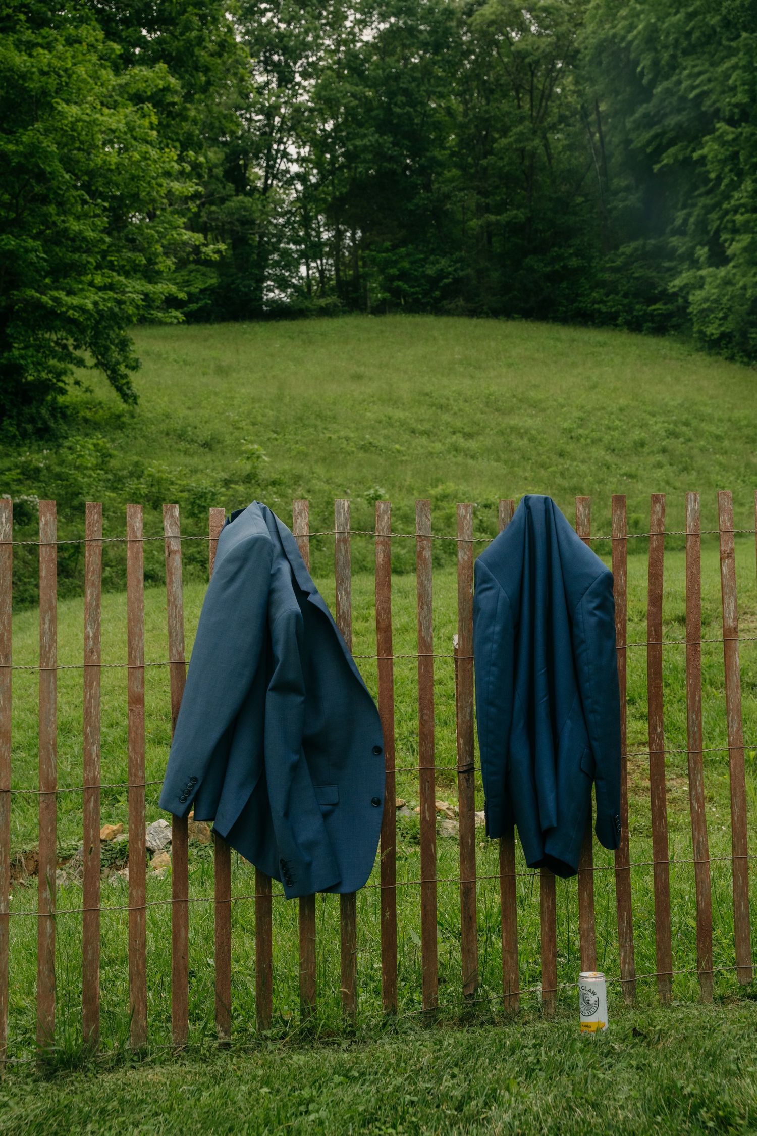 Two blue suit jackets hanging on a rustic wooden fence in a green outdoor countryside setting.