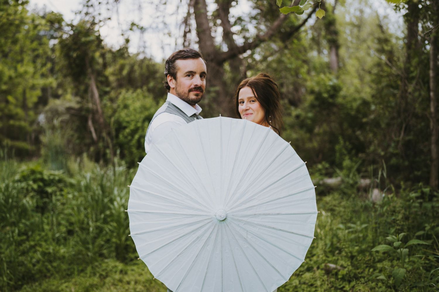 Bride and groom peeking from behind white parasol umbrella in green wooded setting.