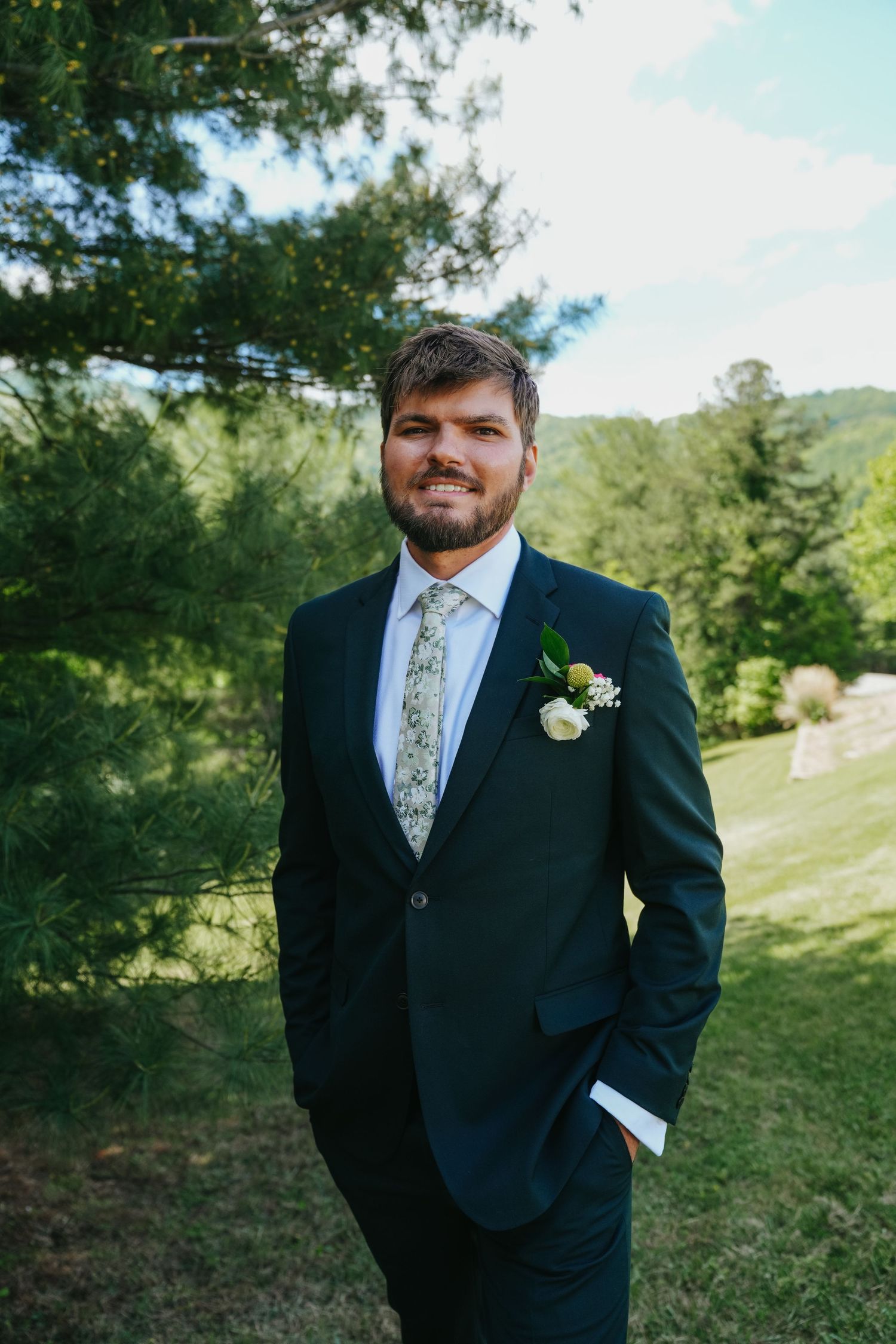 Handsome groom in dark suit with floral boutonniere smiles outdoors near green pine trees.