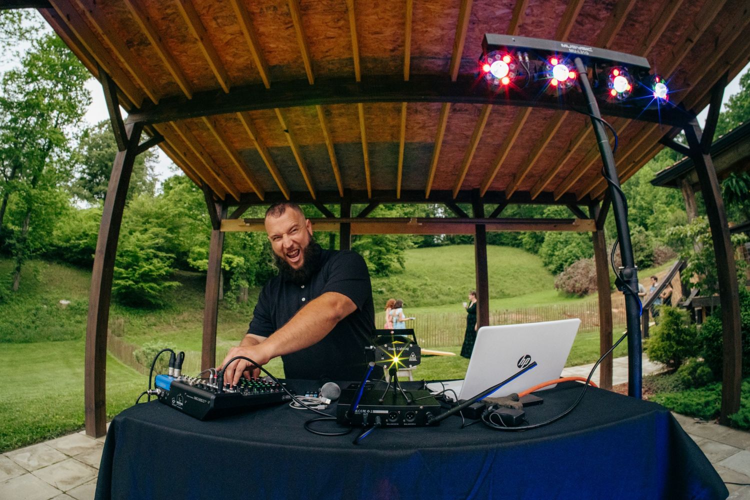Smiling DJ performs at an outdoor wedding reception under a wooden gazebo surrounded by lush greenery.