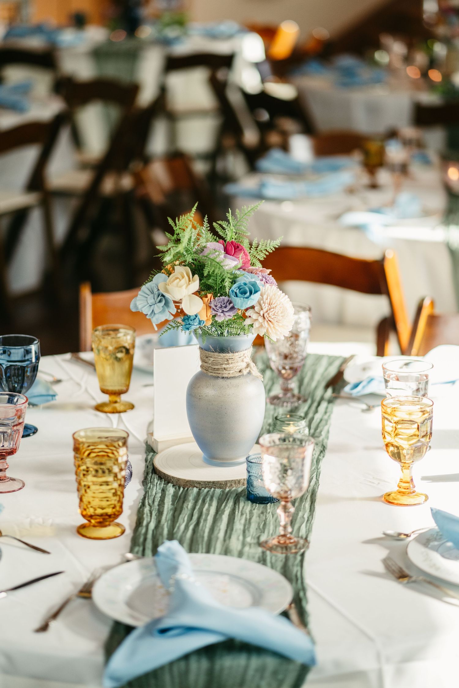 Wedding reception table with gray vase centerpiece, mixed colorful goblets, and blue runner.