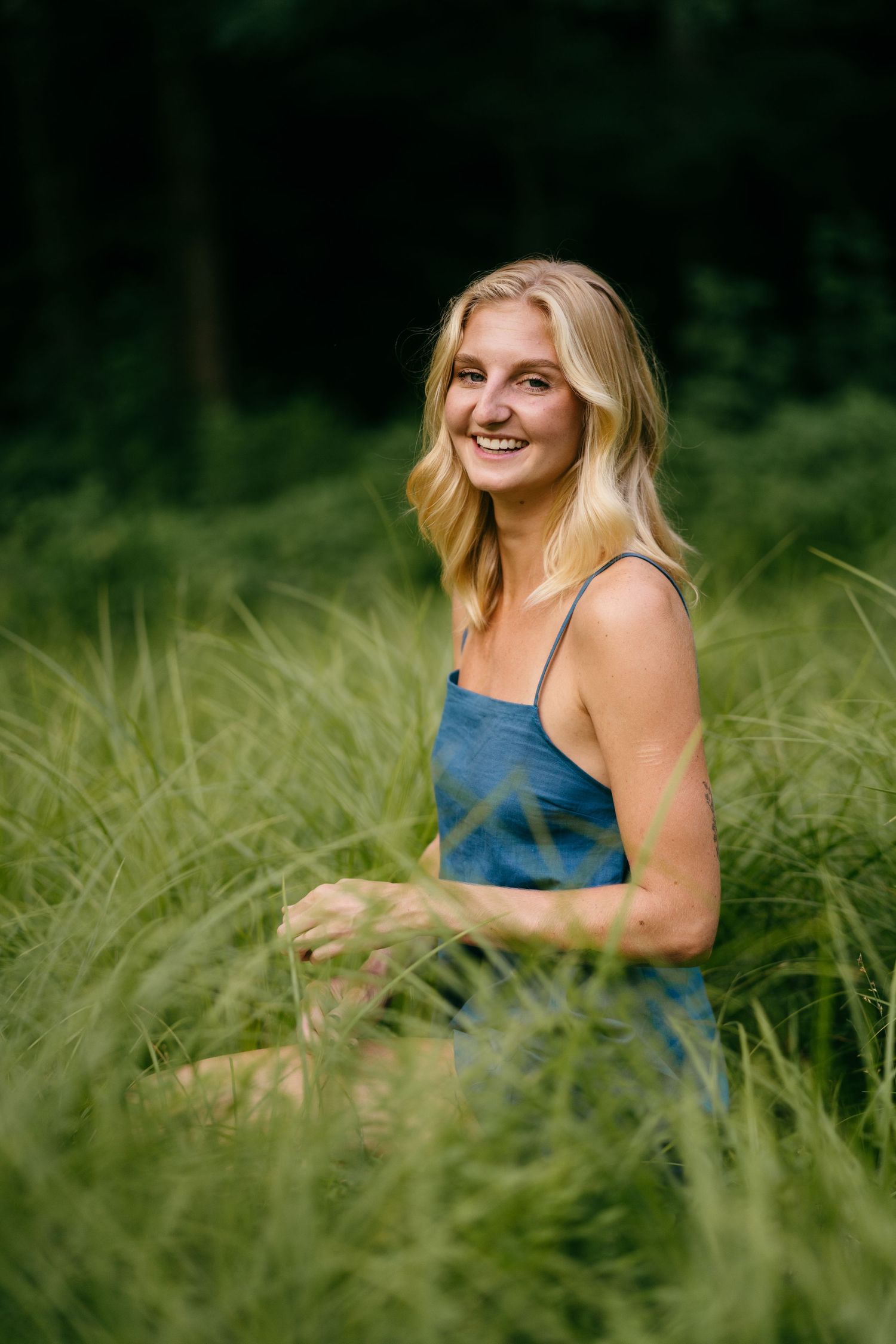 Smiling blonde woman in blue sundress sitting in lush green meadow grass with forest background.