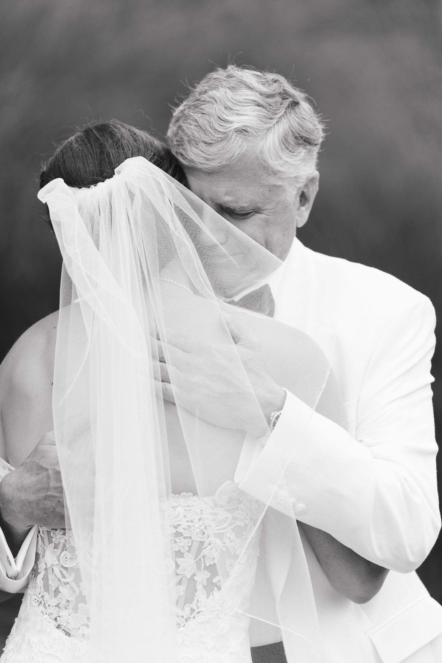 Black and white romantic photo of groom in white suit kissing bride through her flowing wedding veil.