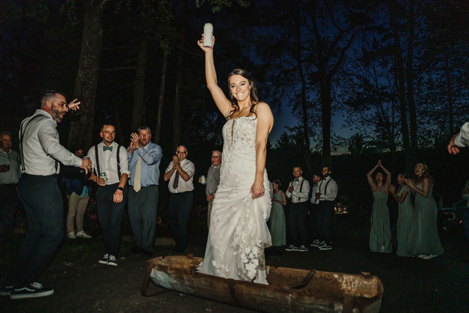 Bride in white gown raises a drink while balancing on a log during a lively outdoor wedding reception at dusk.