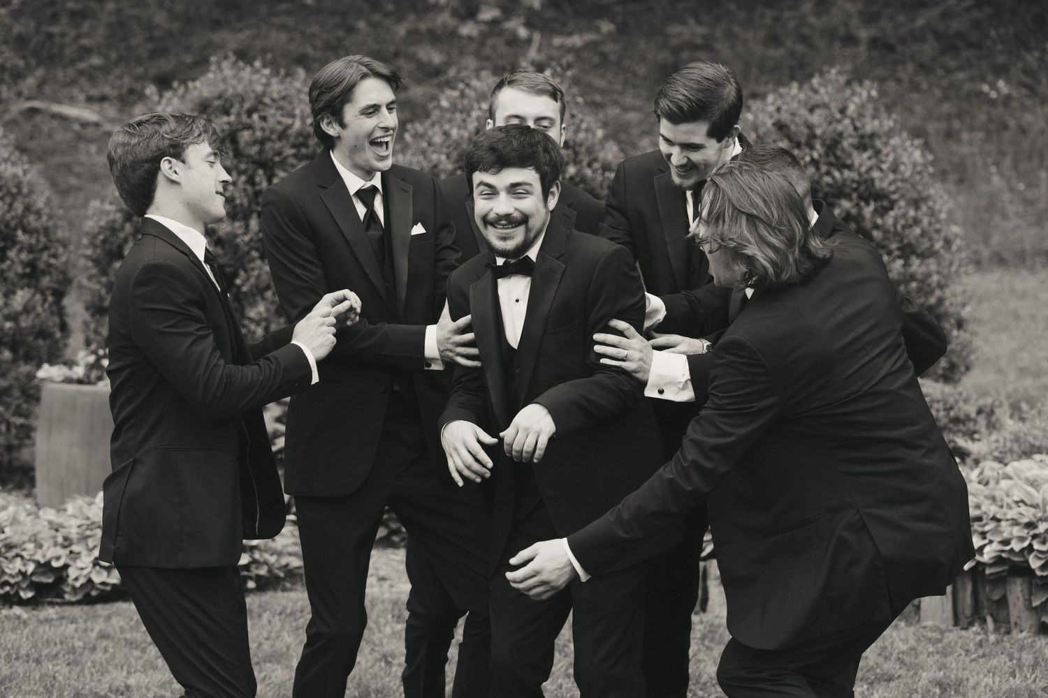 Black and white photo of groom laughing with groomsmen in tuxedos outdoors.
