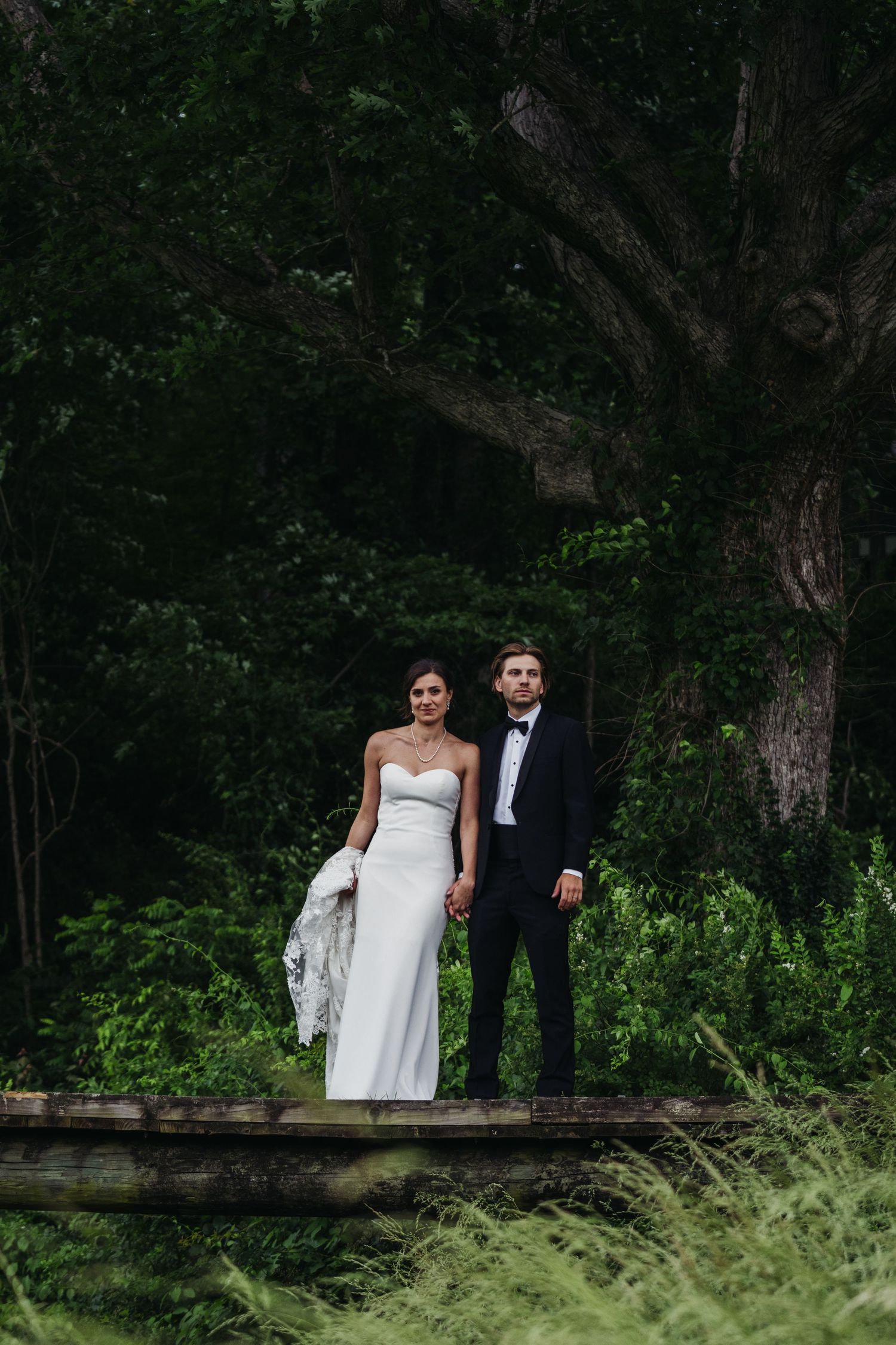 A bride in a white strapless gown and groom in a black tuxedo stand together on a bridge in a forest.