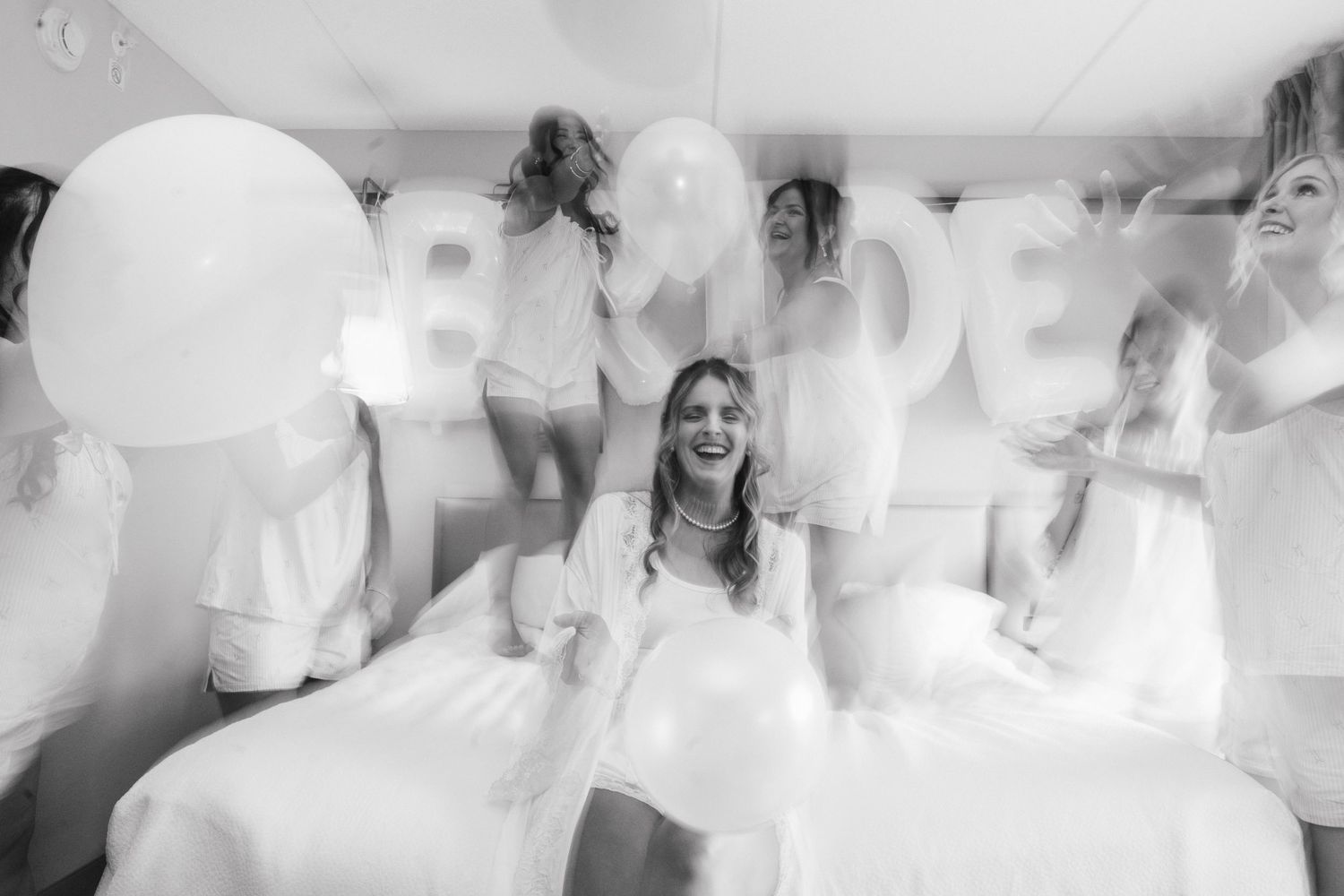 Black and white photo of a bride surrounded by bridesmaids holding white balloons spelling BRIDE on a bed.