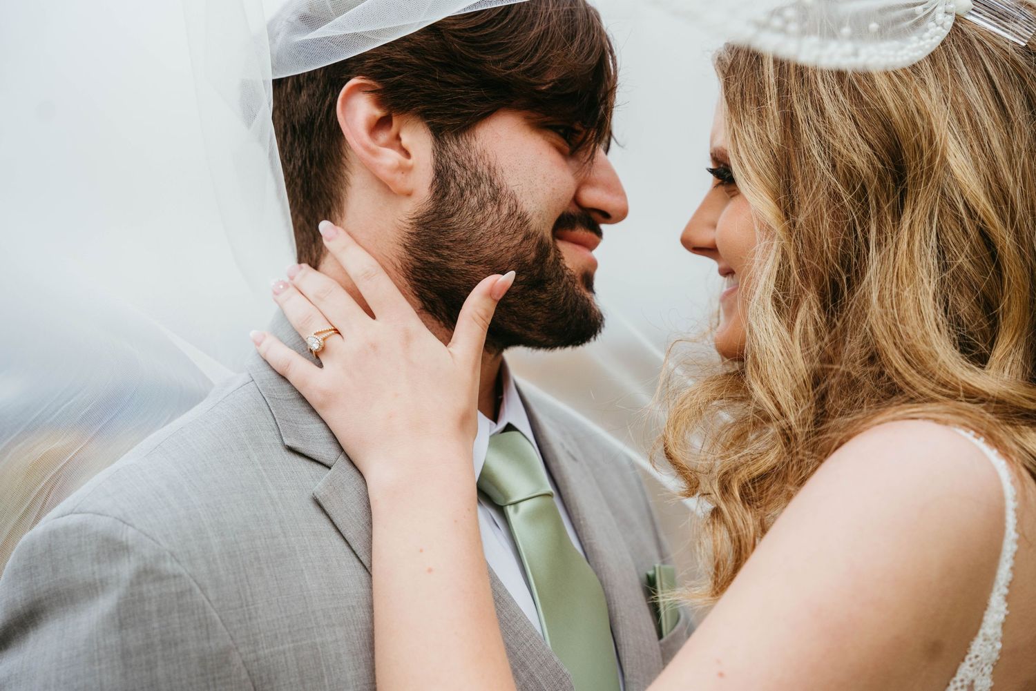Smiling bride and bearded groom share intimate moment under veil, groom wearing gray suit and kippah.