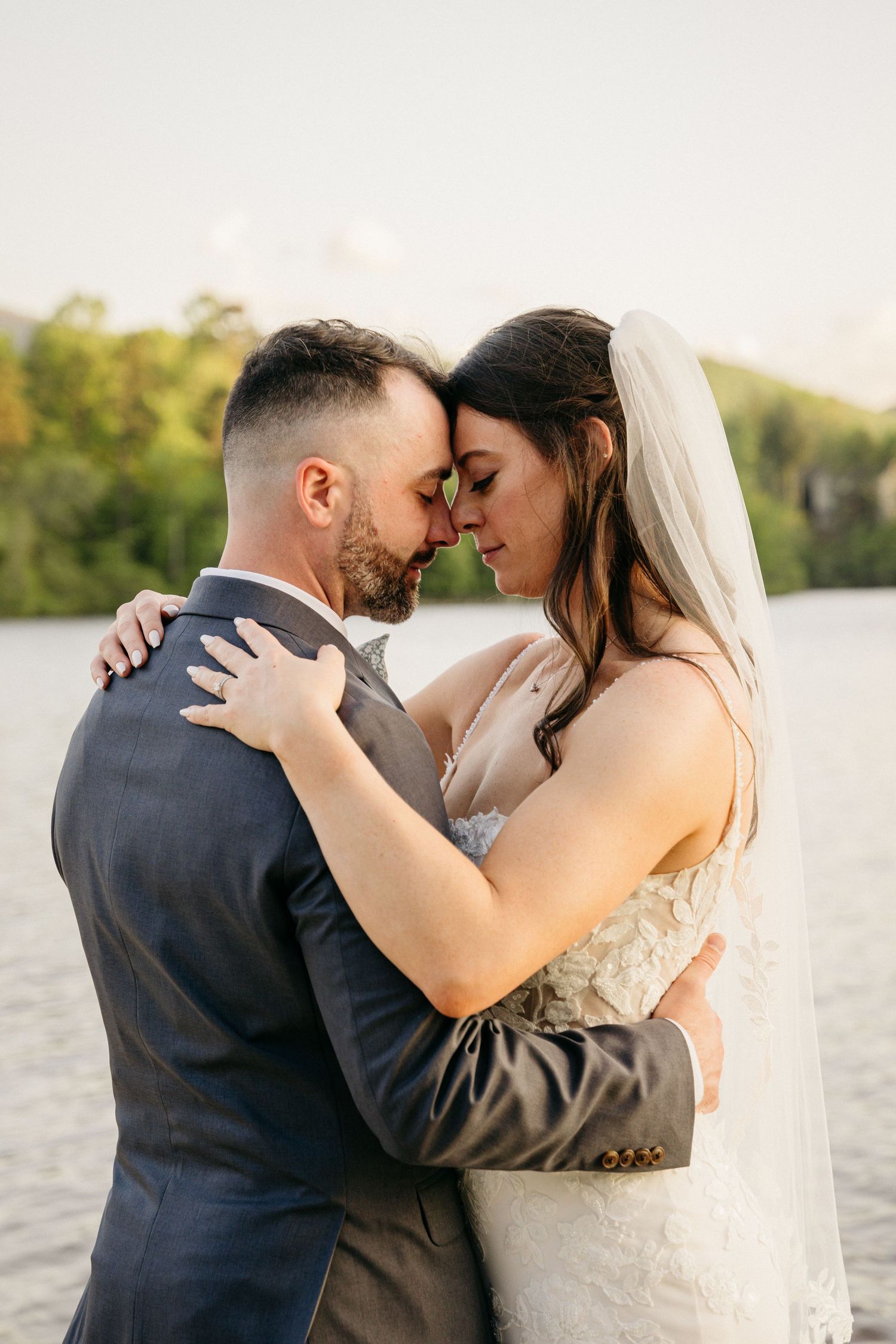 Bride and groom touching foreheads in a romantic embrace by a sunlit lake.