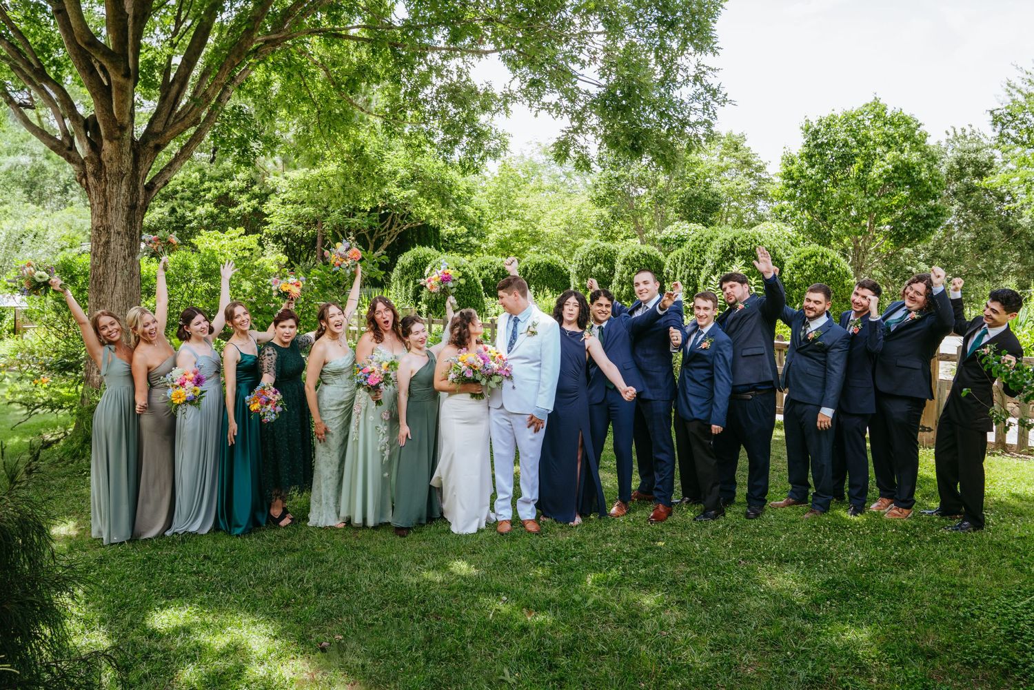 Large wedding party celebrating outdoors under a tree with bridesmaids in green and groomsmen in navy.