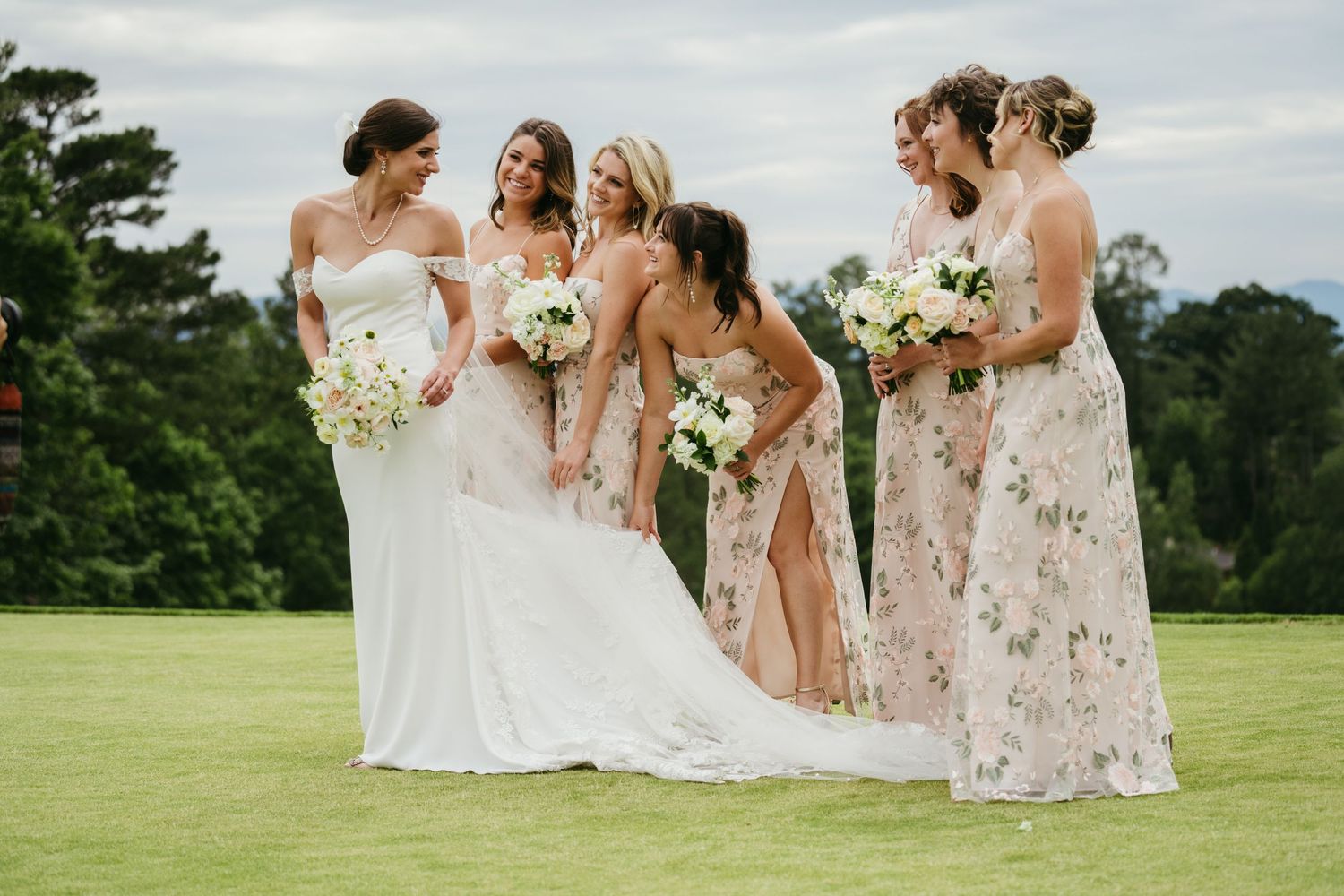 Bride with bridesmaids in floral dresses holding white bouquets on green lawn at wedding.