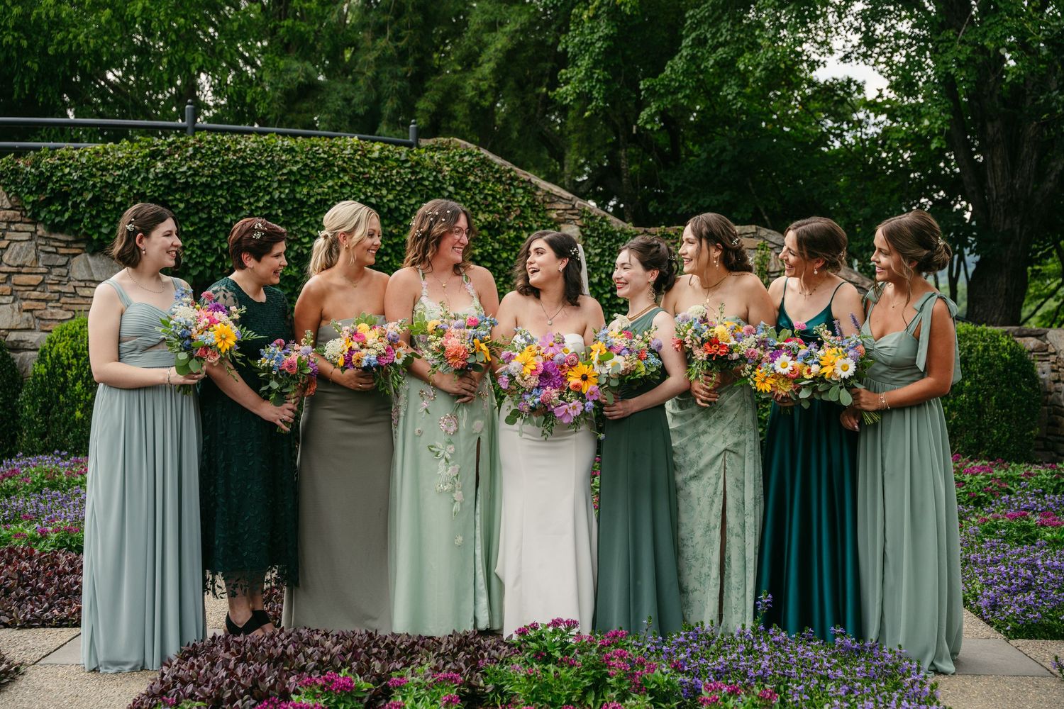 Bride with bridesmaids in mismatched sage and green gowns holding colorful bouquets in garden.