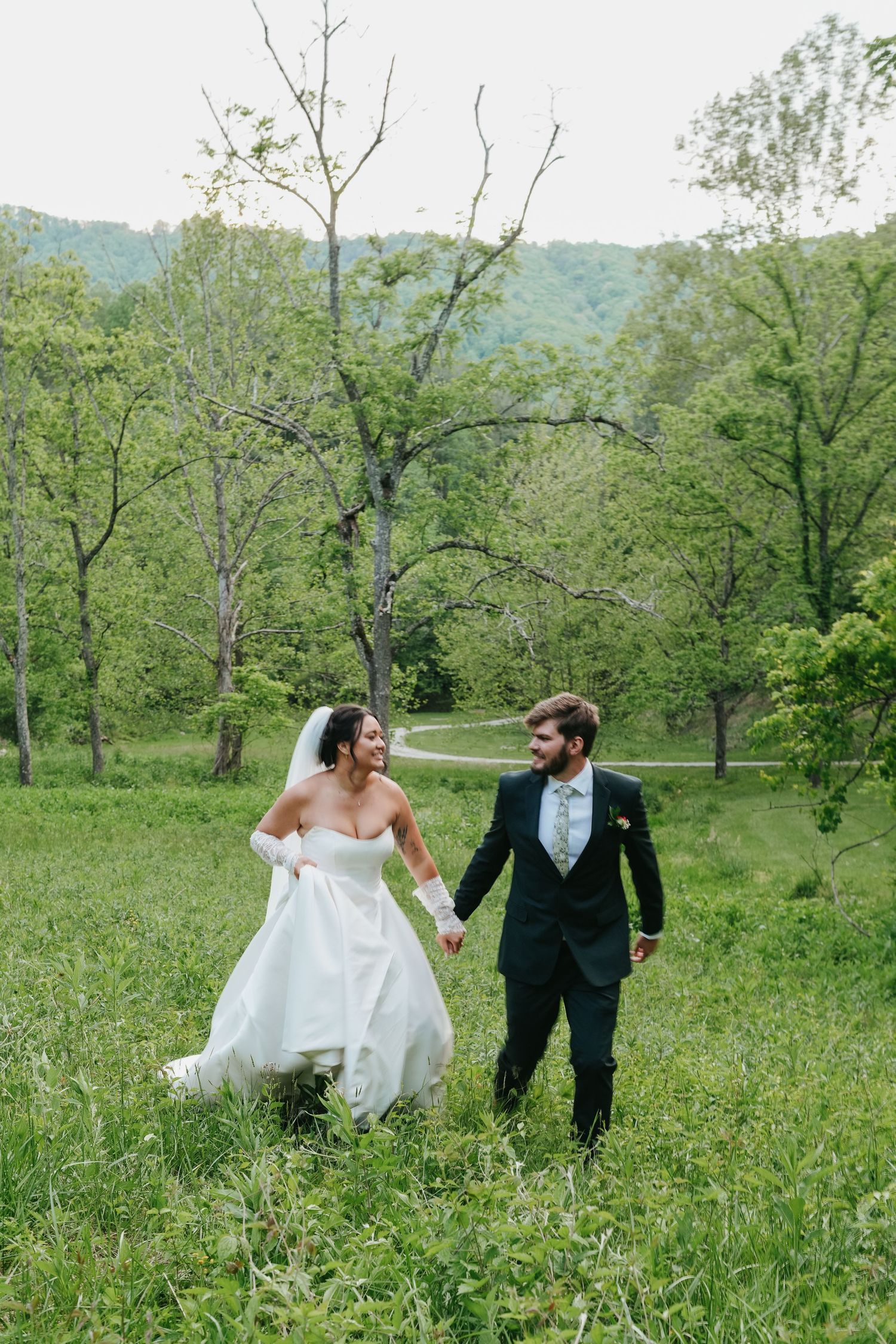 Joyful bride and groom walk hand in hand through a lush green meadow with rolling hills.