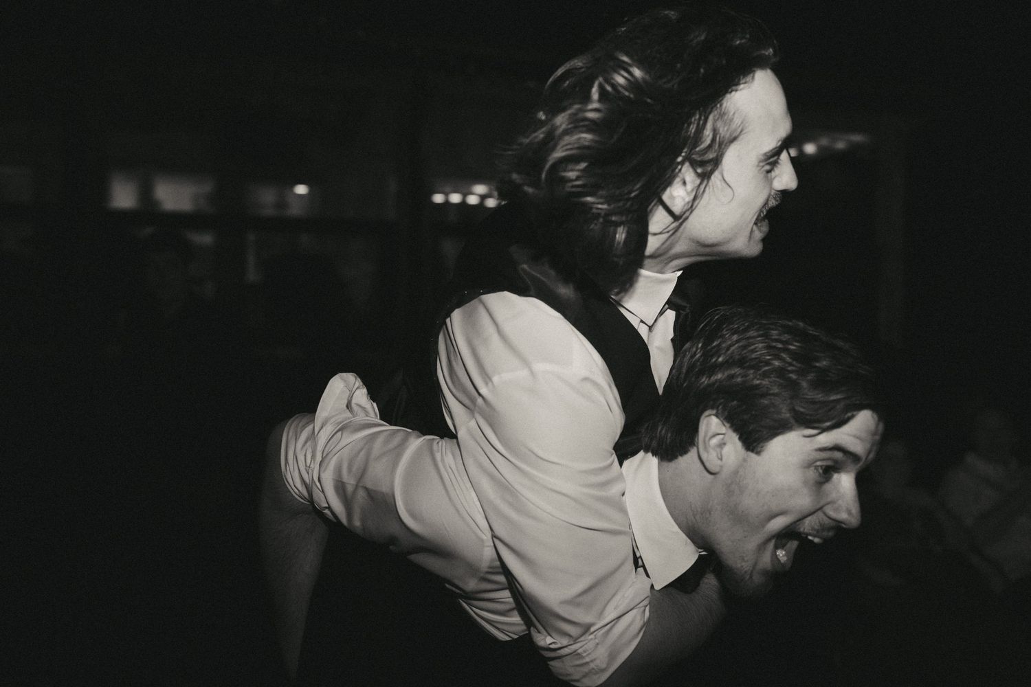 Black and white photo of a groomsman giving another a piggyback ride at a lively wedding reception.