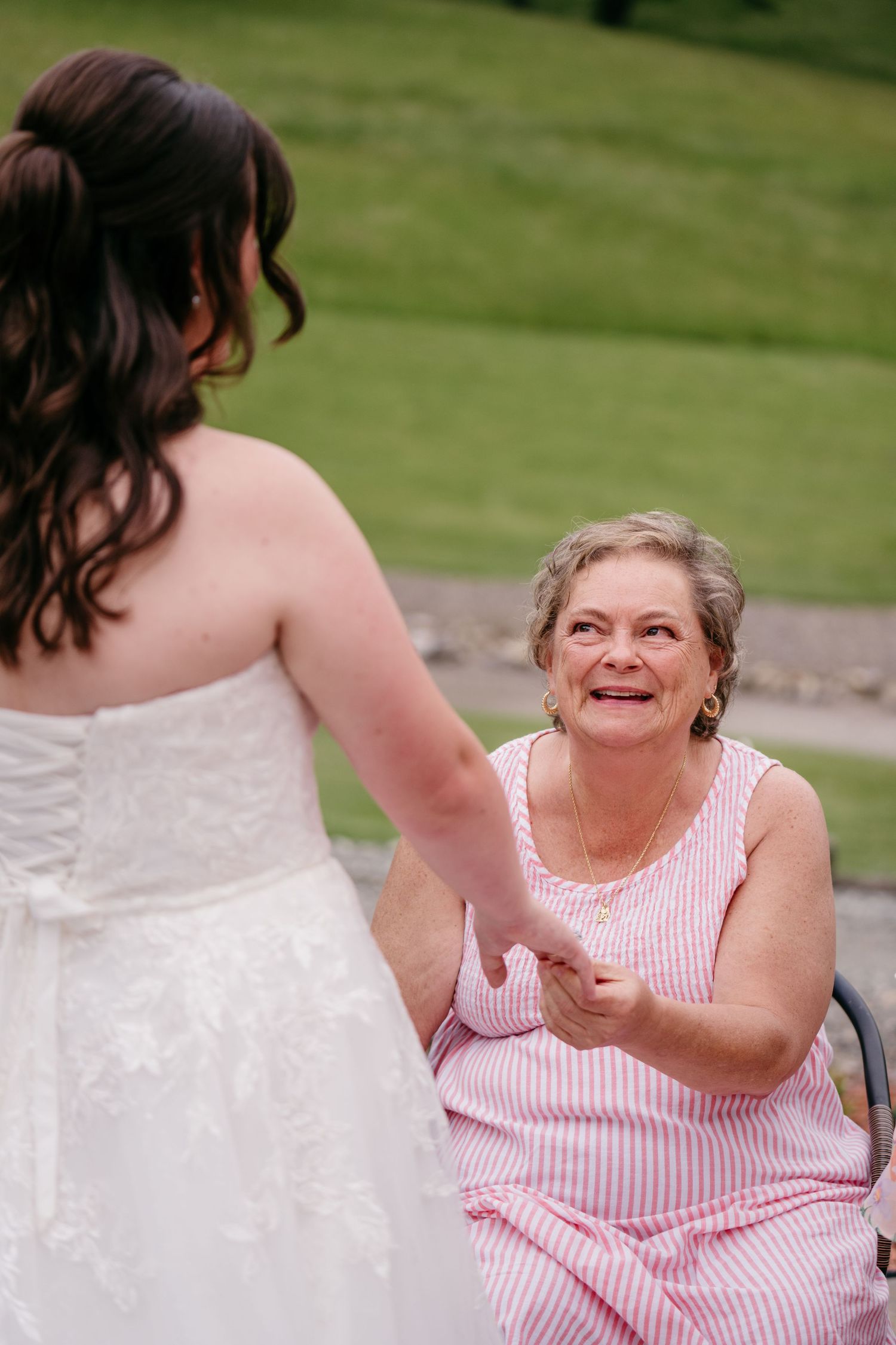 Emotional grandmother in pink dress holds hands with bride in white lace gown at outdoor wedding.