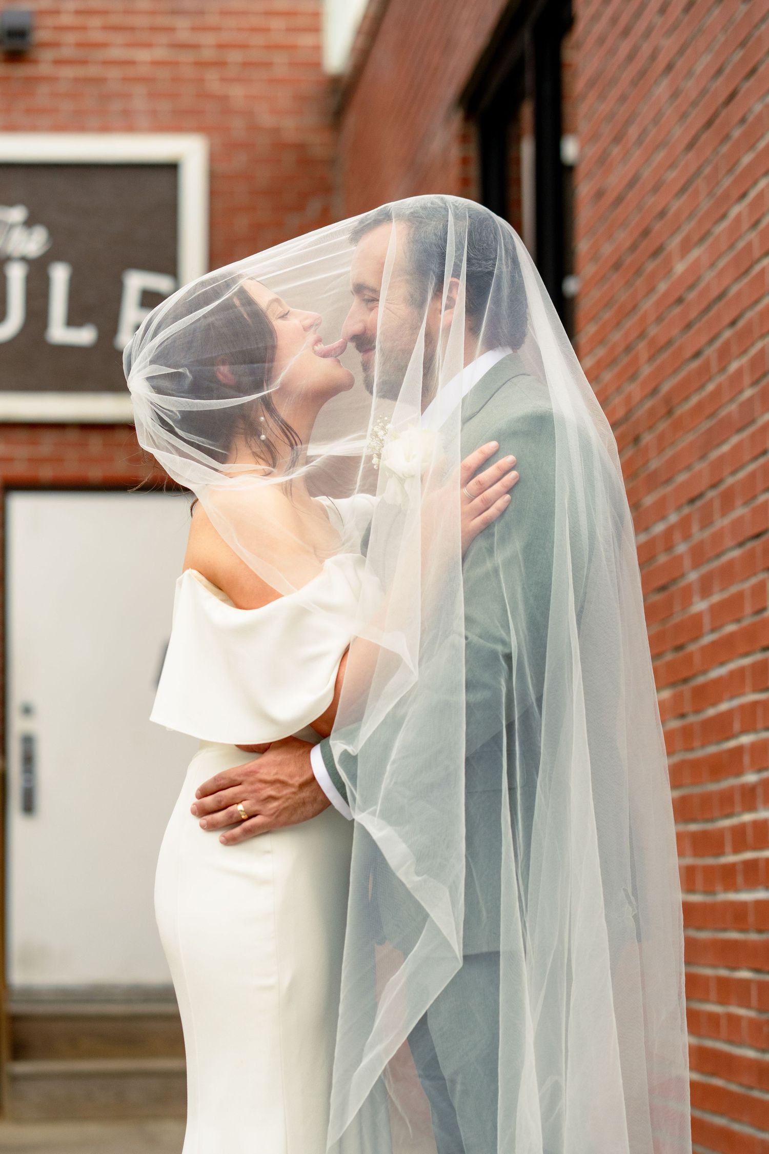 Bride and groom share intimate moment under flowing veil outside brick building.