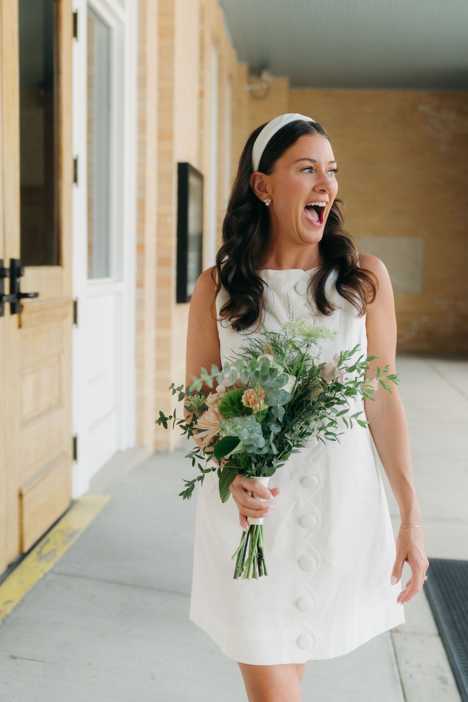 Joyful bride in short white dress with headband laughing while holding green and white bouquet.