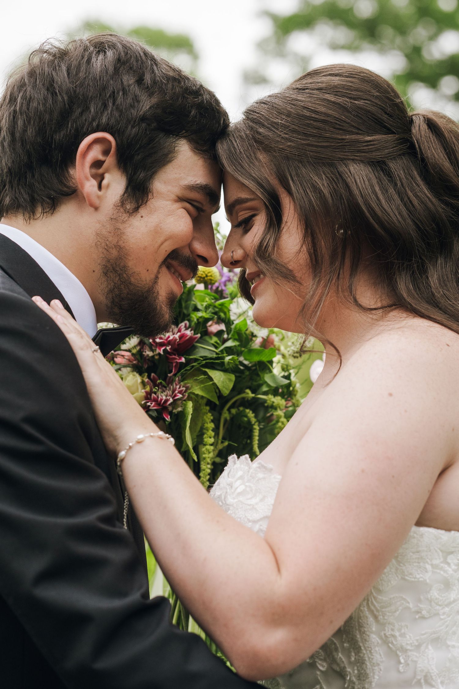 Bride and groom touch foreheads smiling intimately while holding colorful wedding bouquet.