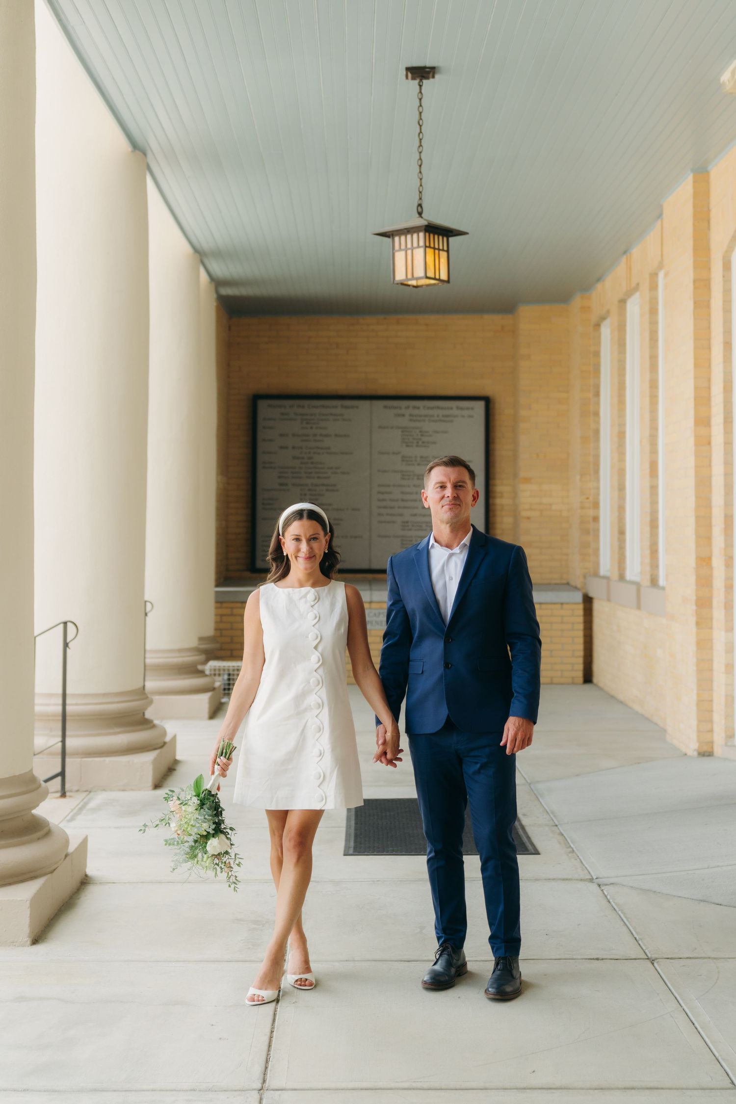 Bride in short white dress and groom in blue suit walk hand in hand through building colonnade.