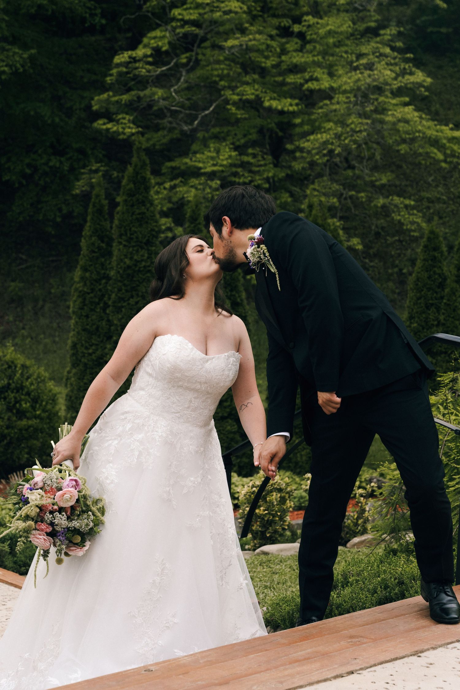Groom in black tuxedo kisses bride in strapless lace gown in lush garden.