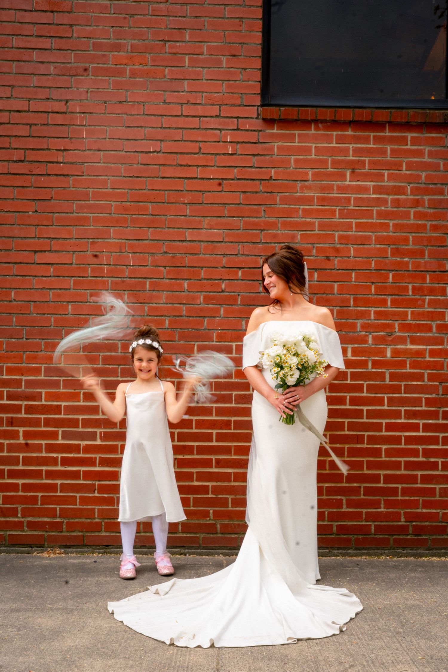 Bride in off-shoulder gown holding bouquet poses with flower girl by brick wall.