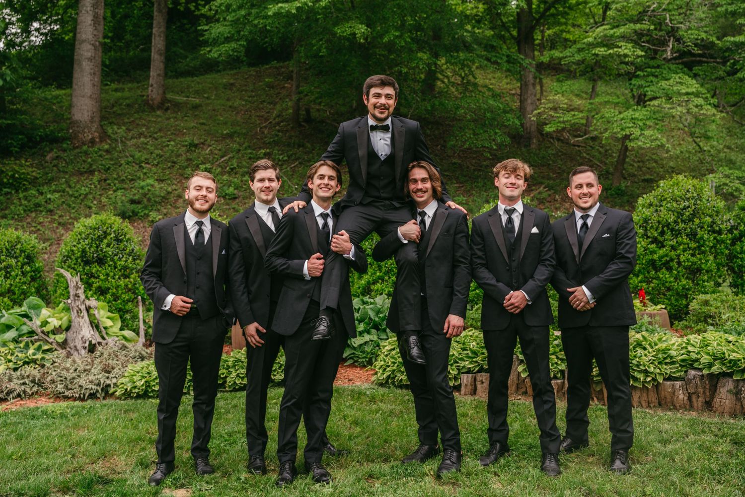 Groom lifted by groomsmen in black suits outdoors at wedding.