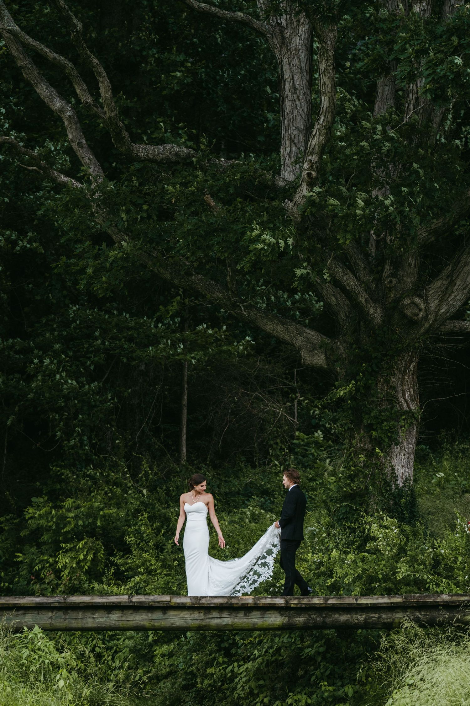 A bride and groom stand facing each other on a rustic bridge surrounded by lush green forest trees.