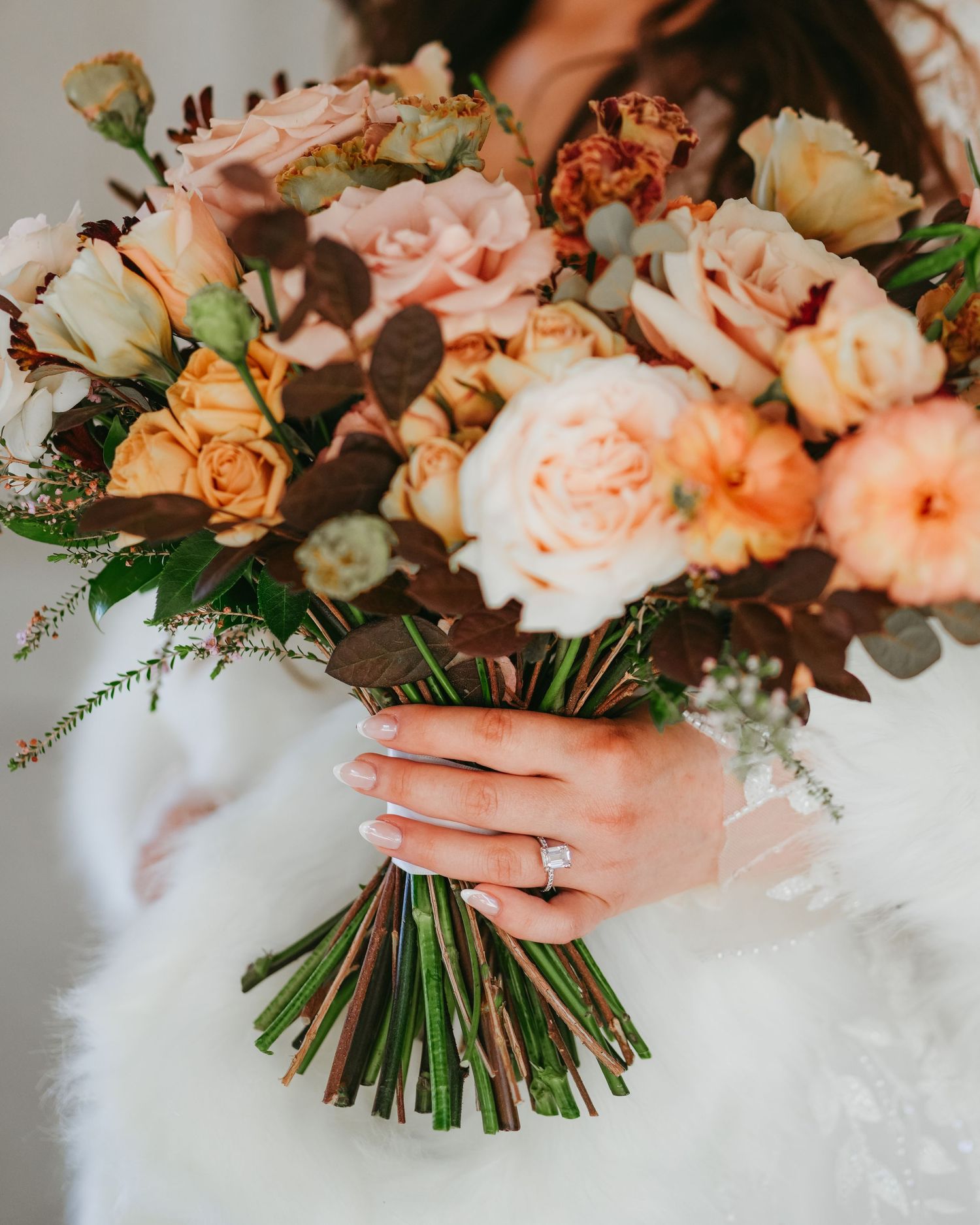 Bride holding a lush autumn wedding bouquet with peach, blush, and rust roses, featuring a diamond engagement ring.
