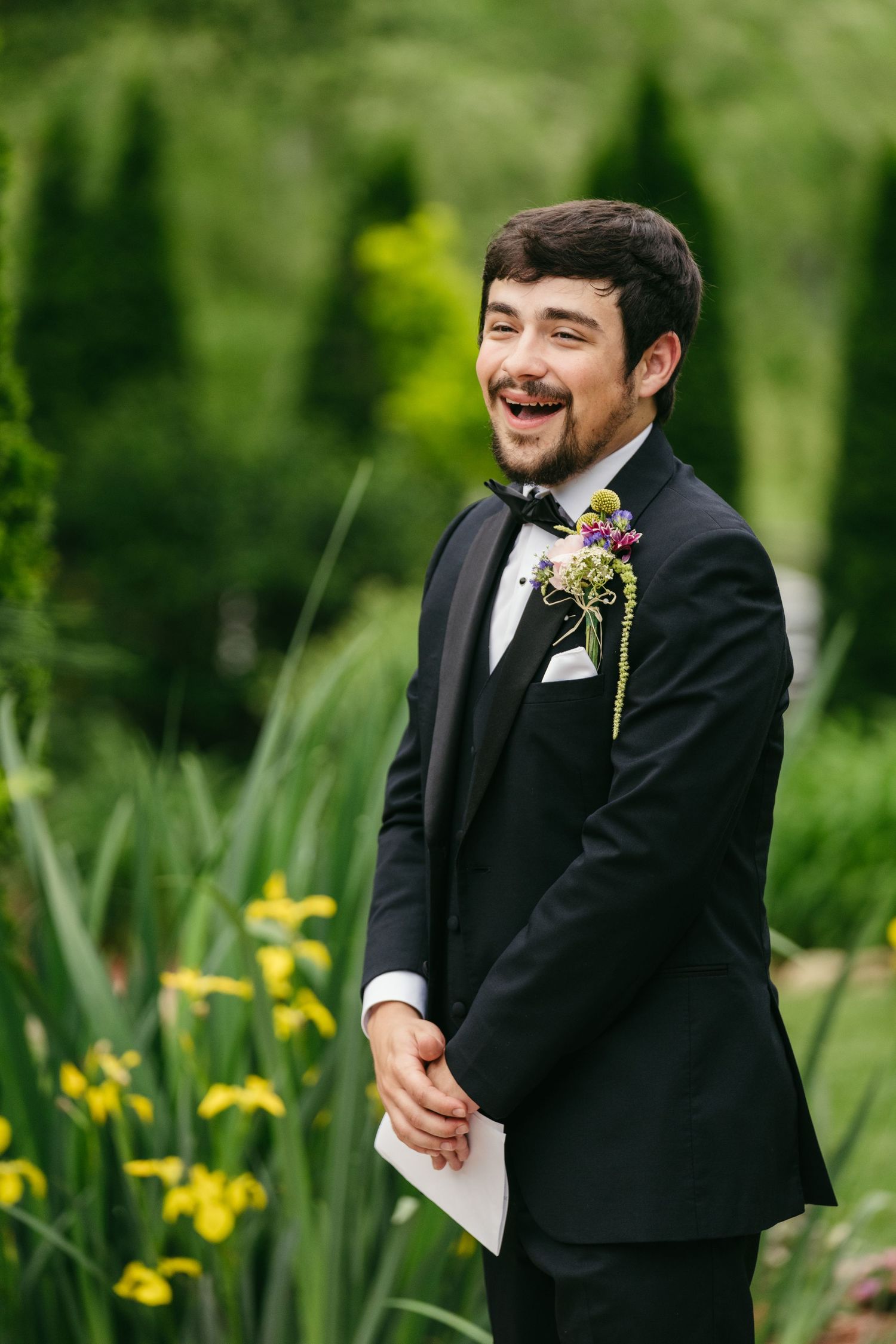 Smiling groom in black tuxedo with boutonniere holds vows in lush garden setting.