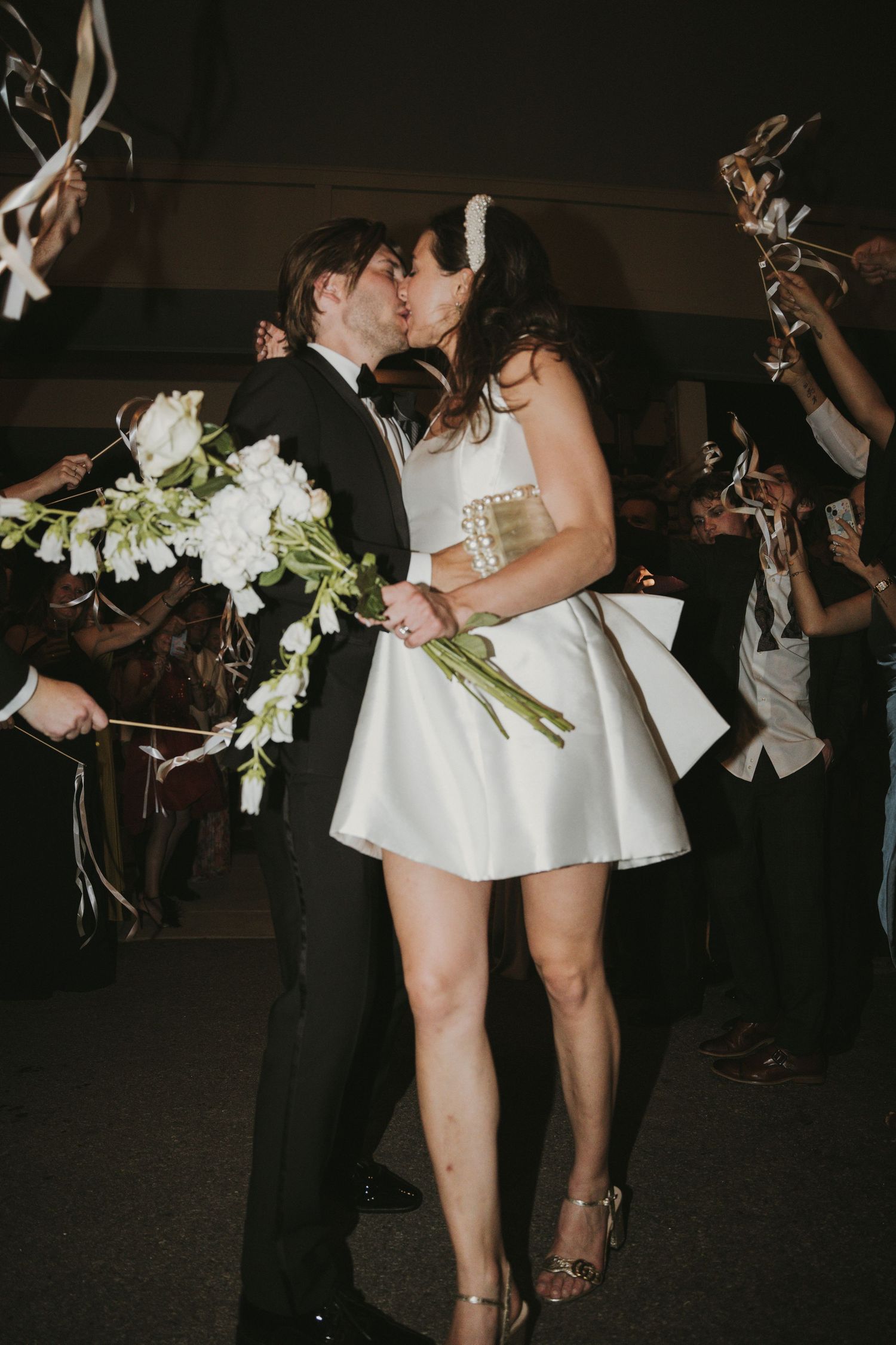 Bride and groom share a kiss during wedding send-off, bride holding white floral bouquet in mini dress.