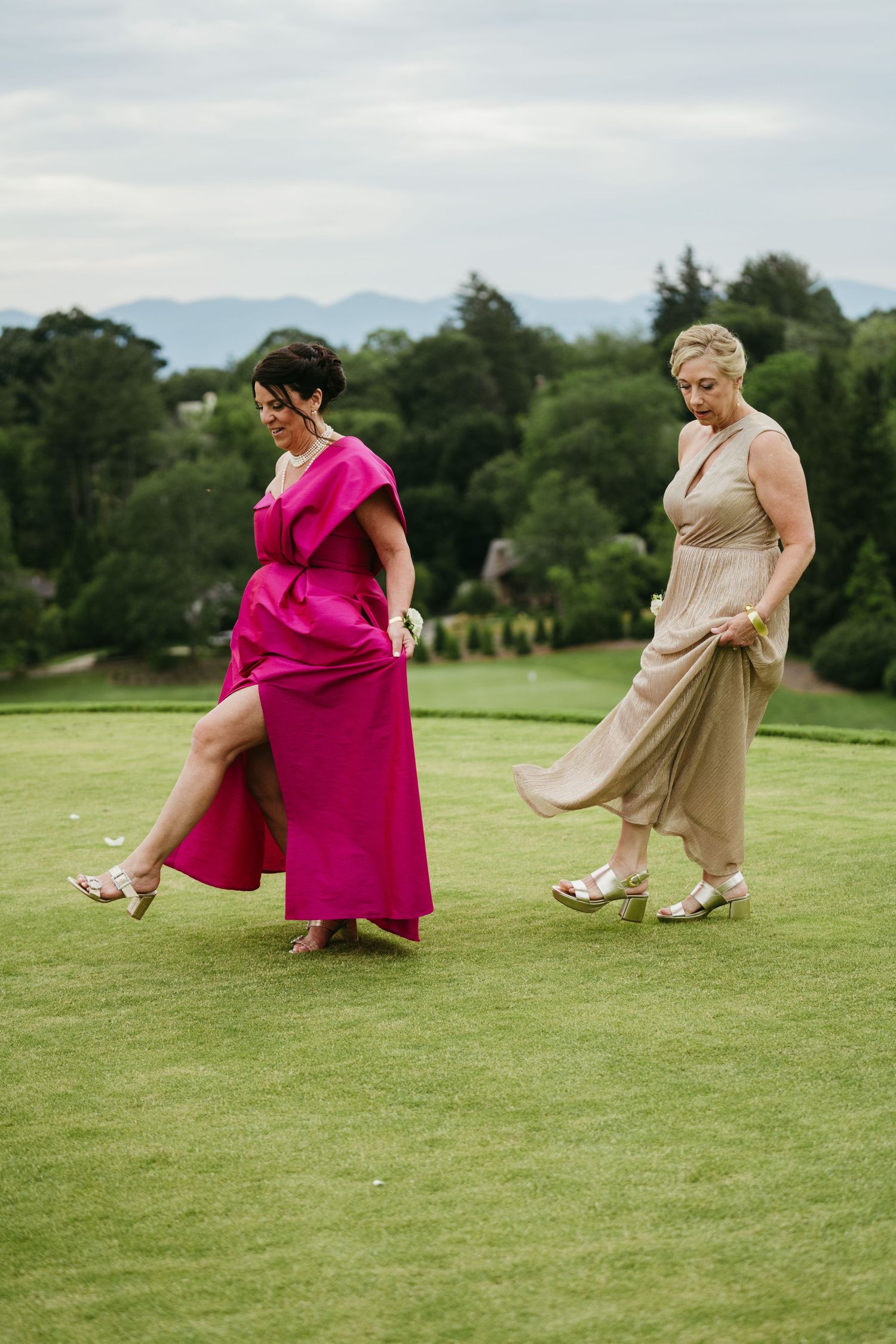 Two elegantly dressed women walk across a lush green lawn, one in a fuchsia gown and one in a champagne dress.
