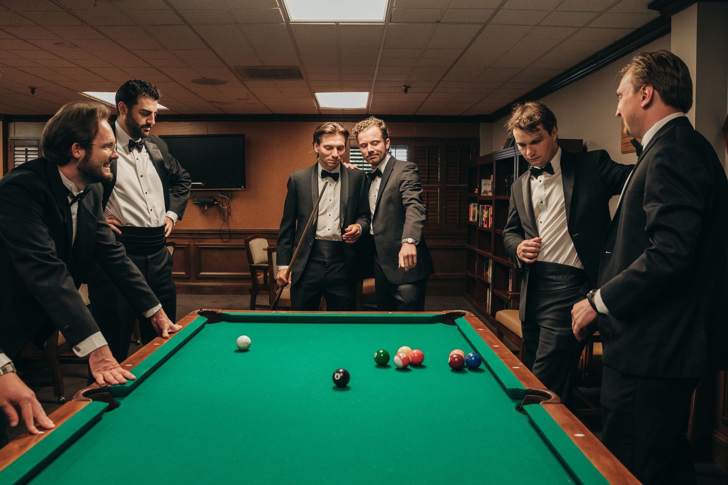 Groomsmen in tuxedos gather around a pool table playing billiards before a wedding celebration.