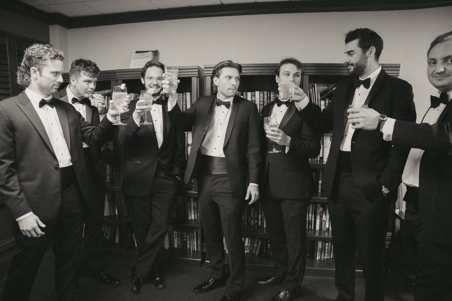 Black and white photo of groomsmen in tuxedos raising glasses in a toast before a wedding ceremony.