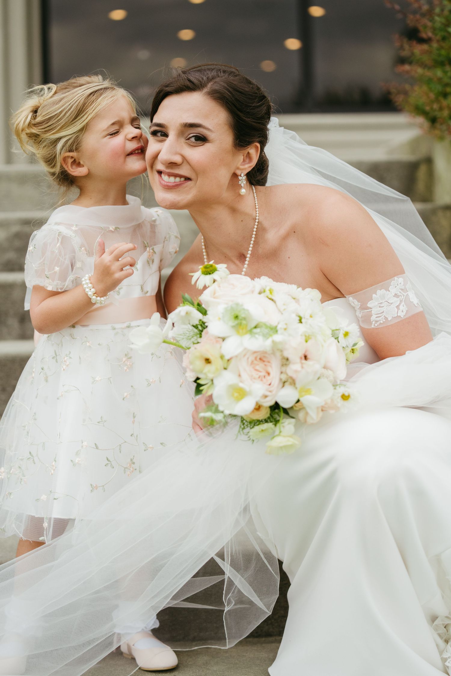 A radiant bride in a white off-shoulder gown holds a bouquet while a young flower girl whispers in her ear.