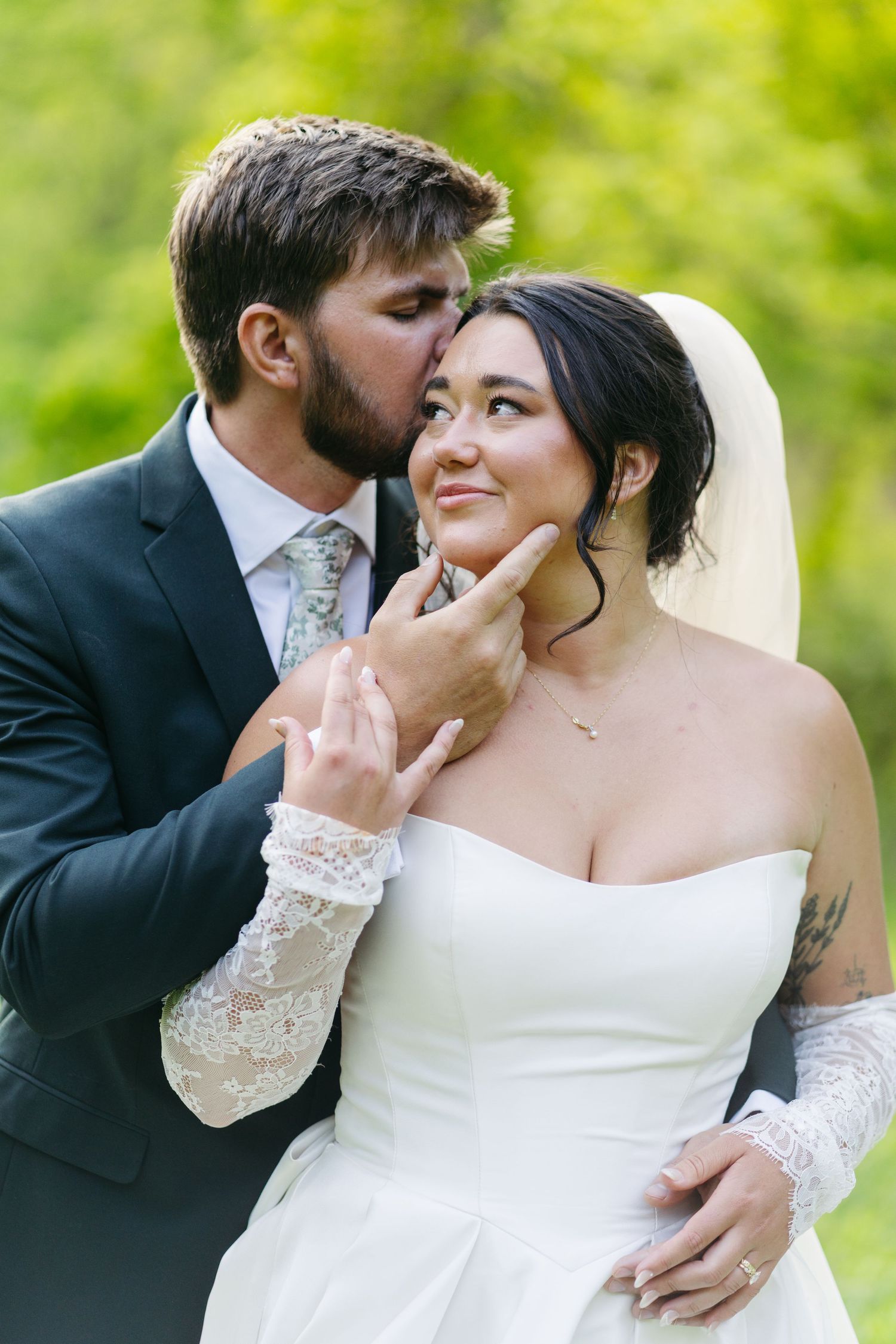 Groom tenderly kisses bride on cheek while holding her chin in a green outdoor setting.