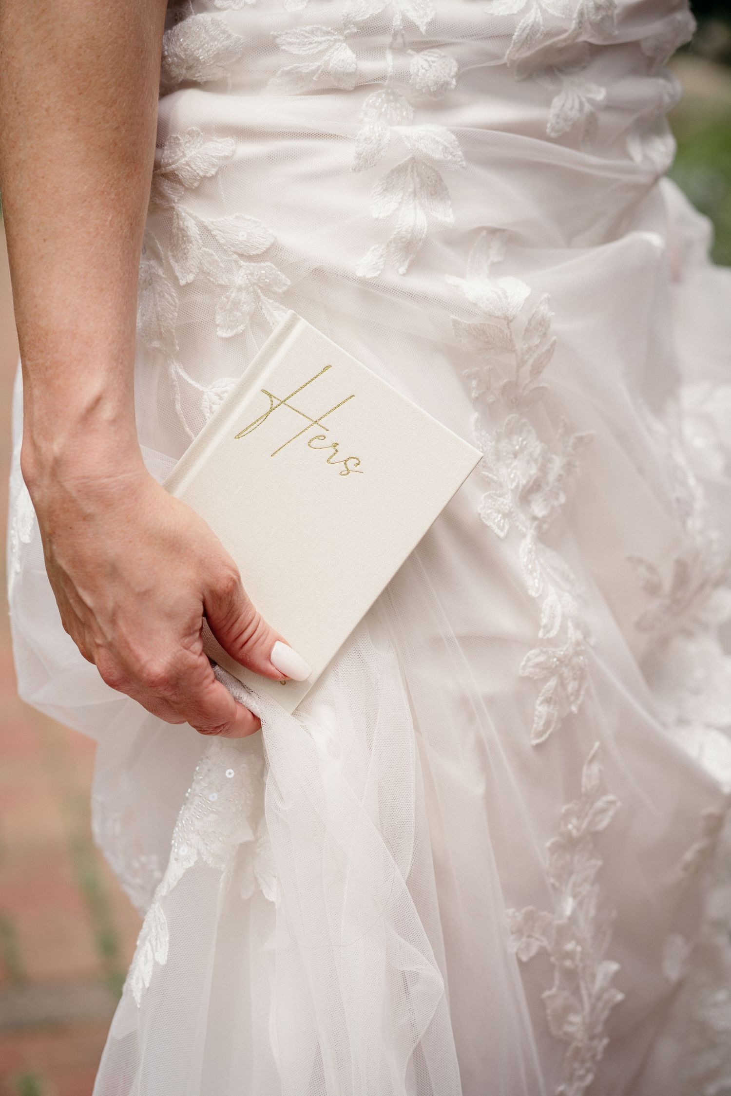 Bride in lace wedding gown holding a white and gold 'Hers' vow book.
