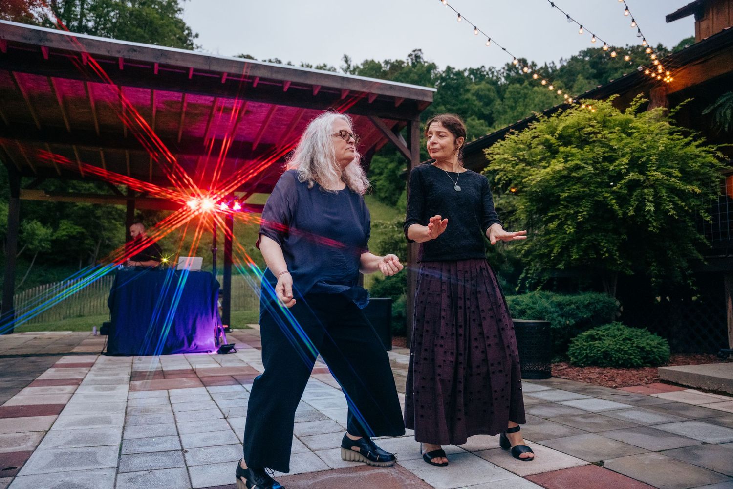 Two women dance joyfully on an outdoor patio illuminated by colorful DJ lights at a wedding reception.