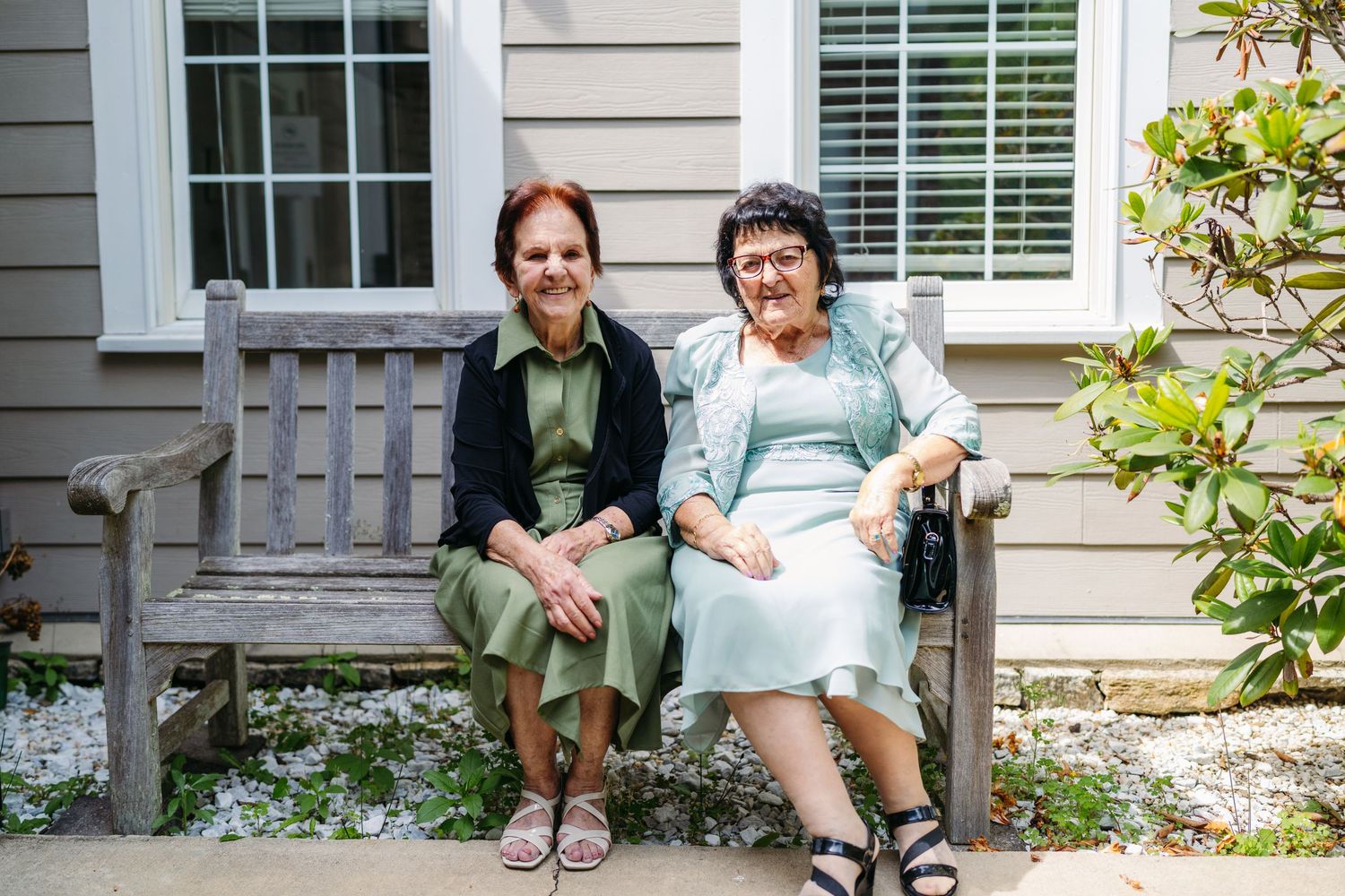 Two elderly women smile while sitting together on a wooden bench outside a house.
