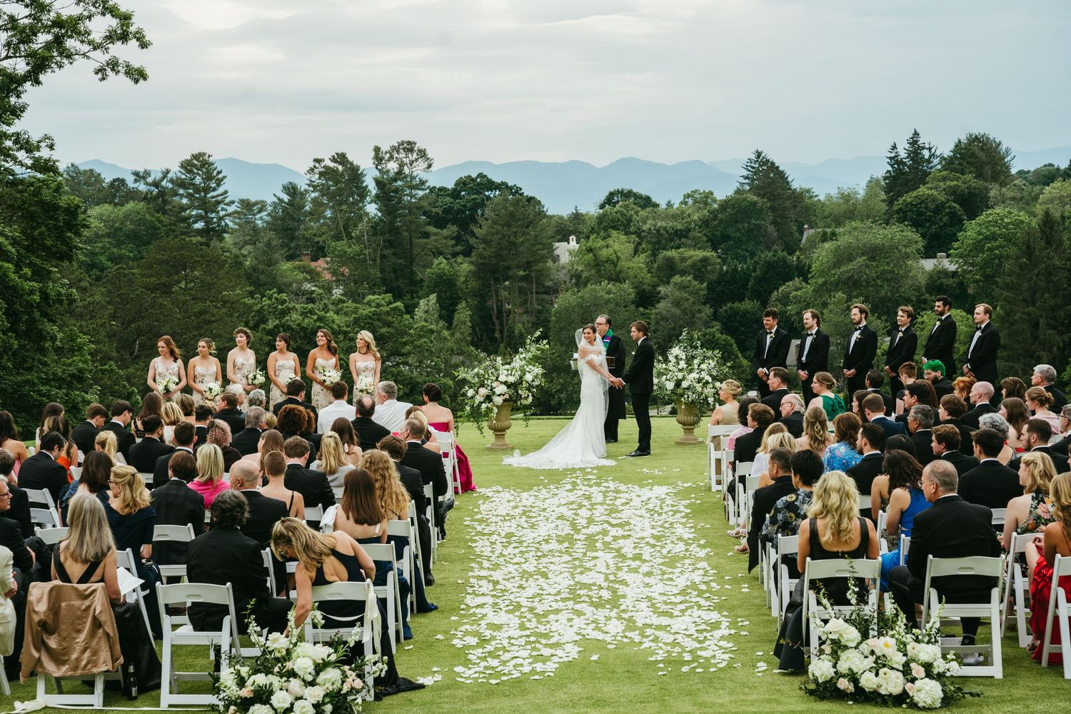 An elegant outdoor wedding ceremony on a lush green lawn with mountain views and white floral aisle decorations.